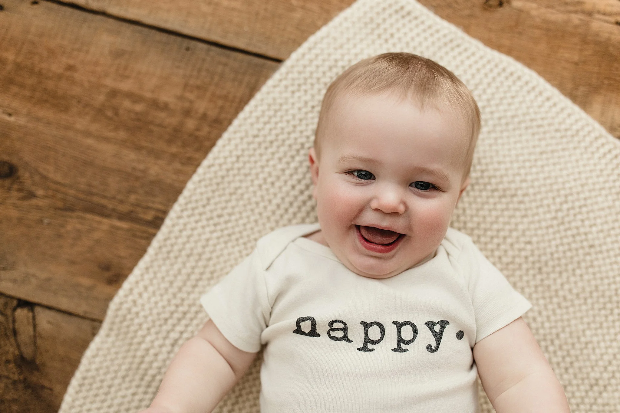 A smiling baby lying on a textured beige blanket on a wooden floor, wearing a white onesie with the word 'happy.' printed on it for one year baby photoshoot in Naperville.