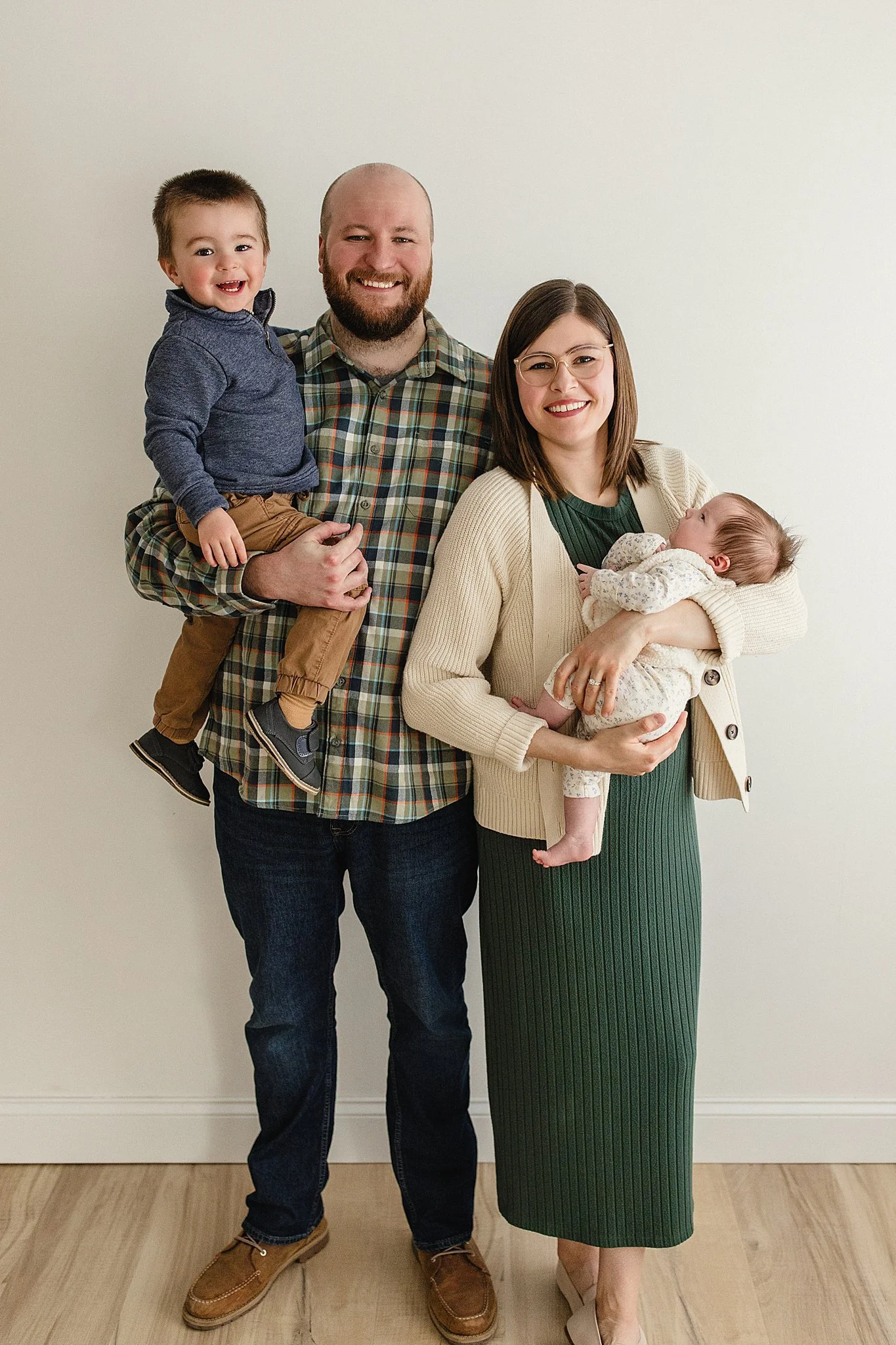 A smiling family of four standing against a plain white wall. The father, with a beard, is holding a young boy with short hair on his left arm. The mother, wearing glasses and a green dress, is holding a baby girl during a studio family photo session