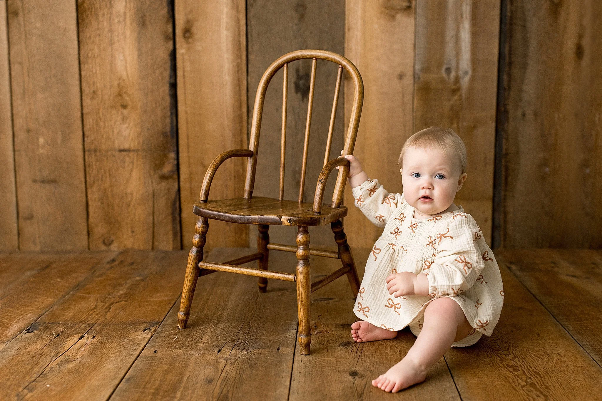 A young child sitting on a wooden floor holding onto the armrest of a wooden chair with a high back. The child has light hair and is wearing a beige dress with orange bow patterns. The background features a wooden wall for baby photos near me.