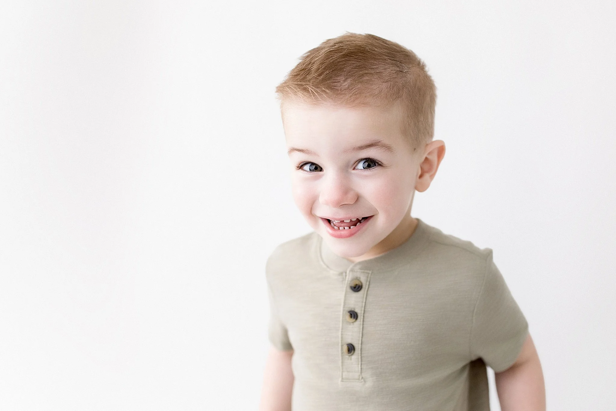 Smiling young boy with light brown hair and blue eyes wearing a beige shirt with buttons, standing against a plain white background for kids photos near me.