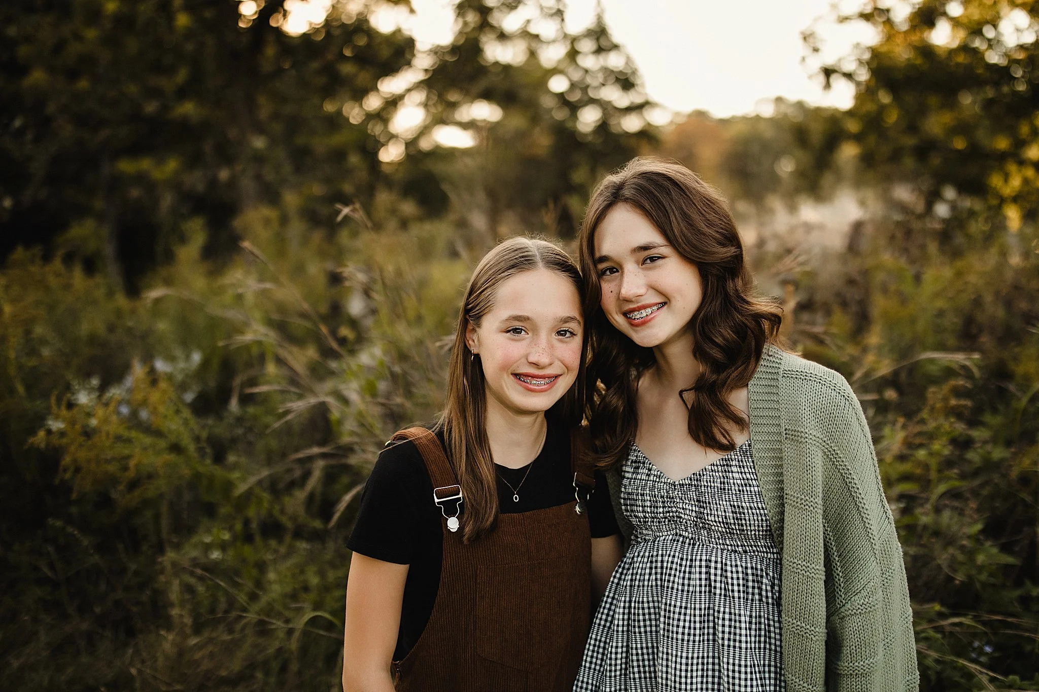 Two teenage girls standing outdoors, smiling at the camera, with trees and foliage in the background during sunset with a Naperville family photographer.