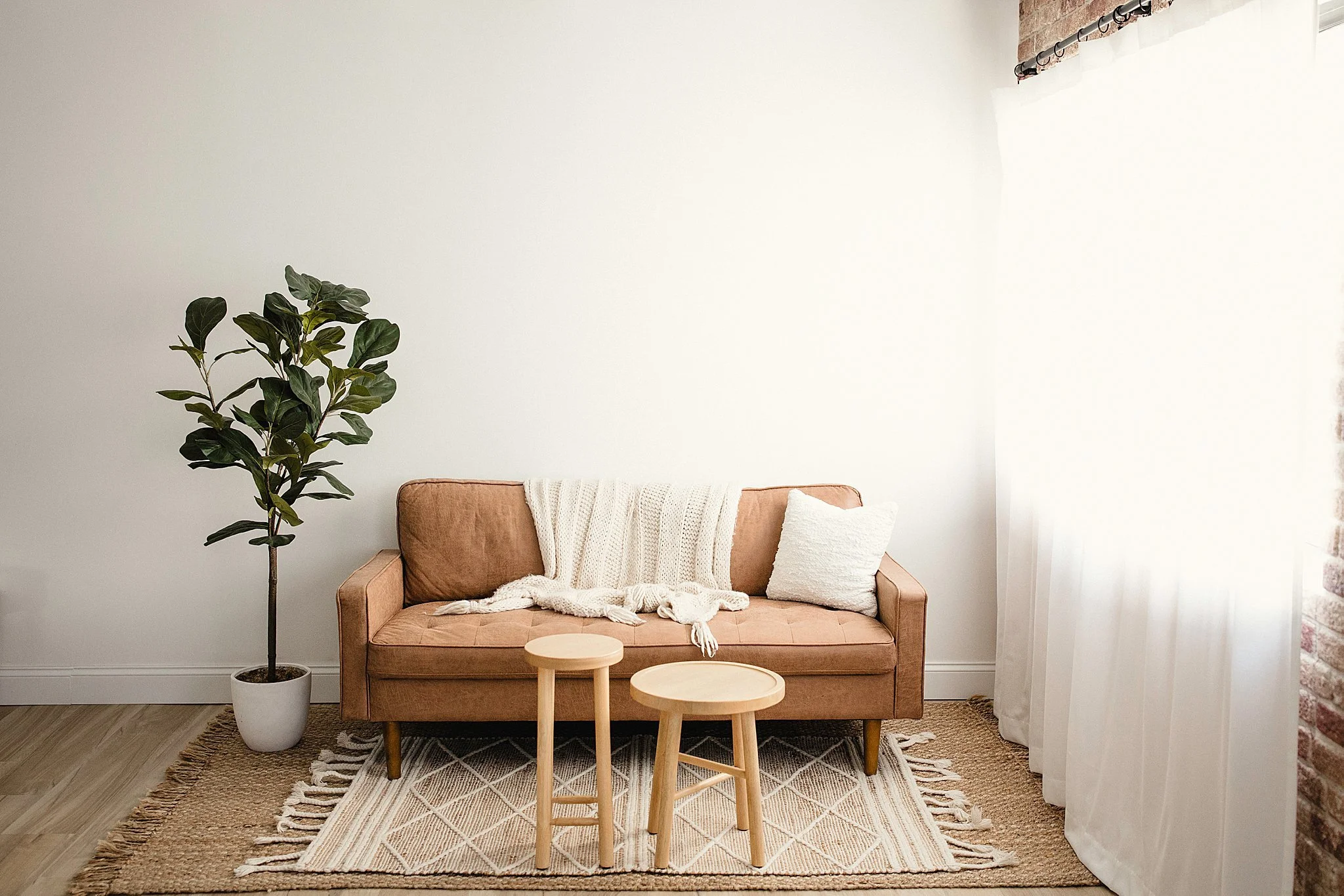 Photo studio room with a brown sofa, a white blanket and pillow, two wooden stools, a potted plant, a woven rug, white curtains, and a partial exposed brick wall in the Ally and B Photography studio in Naperville.