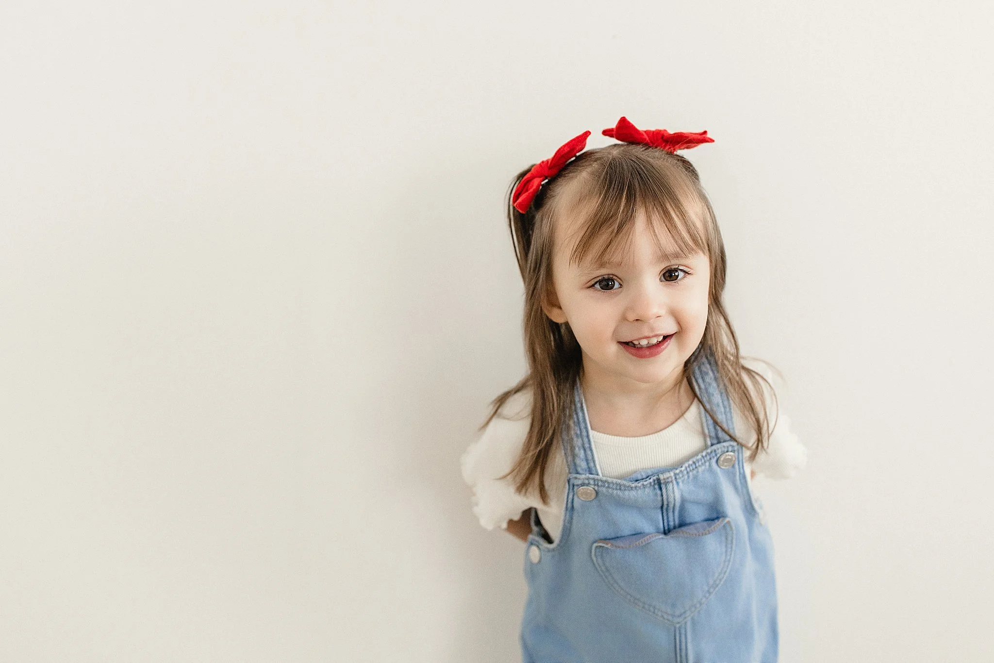A young girl with brown hair wearing a white shirt, denim overalls with a heart pocket, and a red bow in her hair, standing against a plain light-colored background for kids modeling near me.