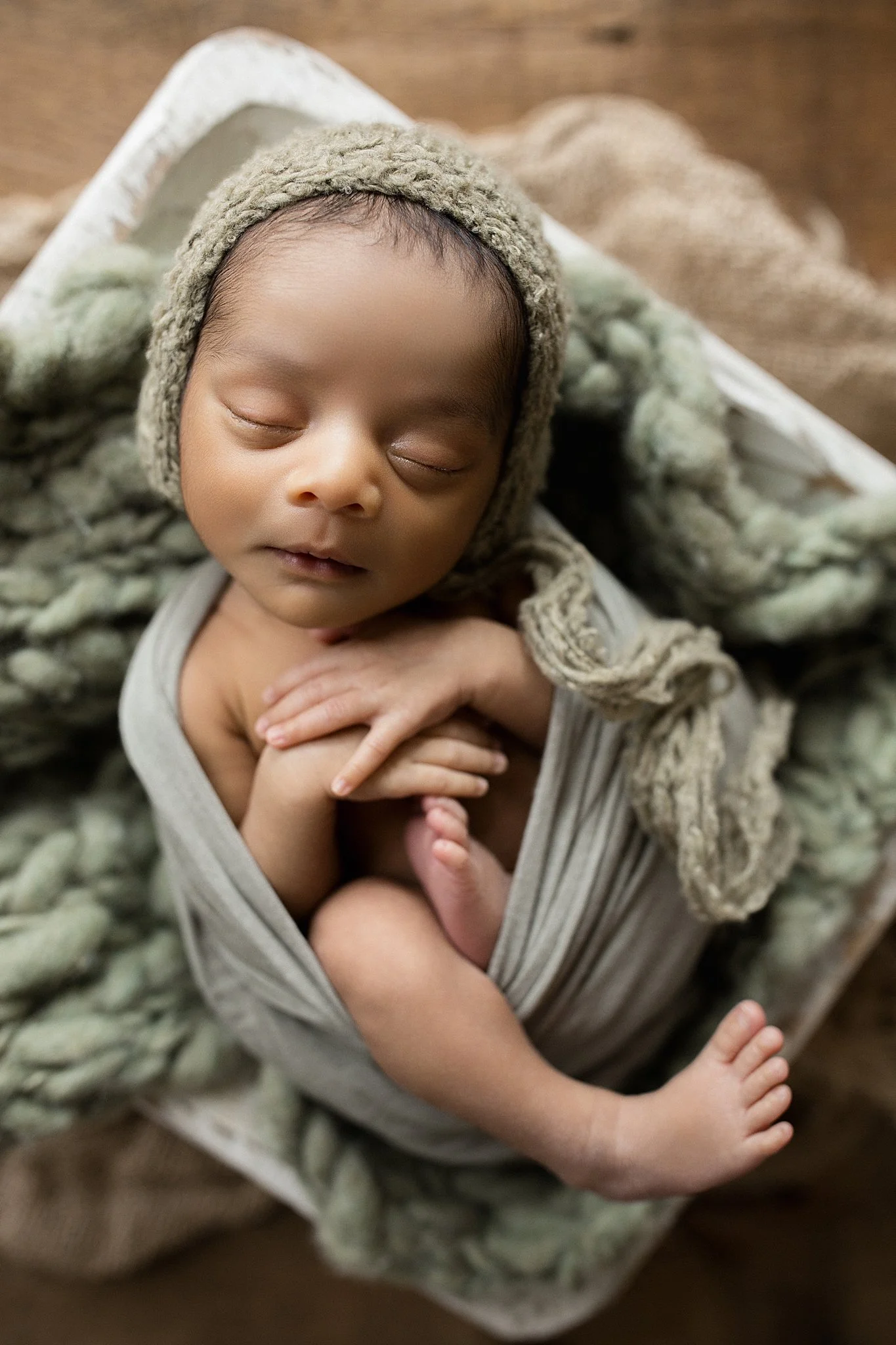 A sleeping baby wrapped in a gray swaddle, wearing a knit hat, laying on a textured blanket with a wooden floor background during posed newborn photos with marie lopez in naperville, IL.