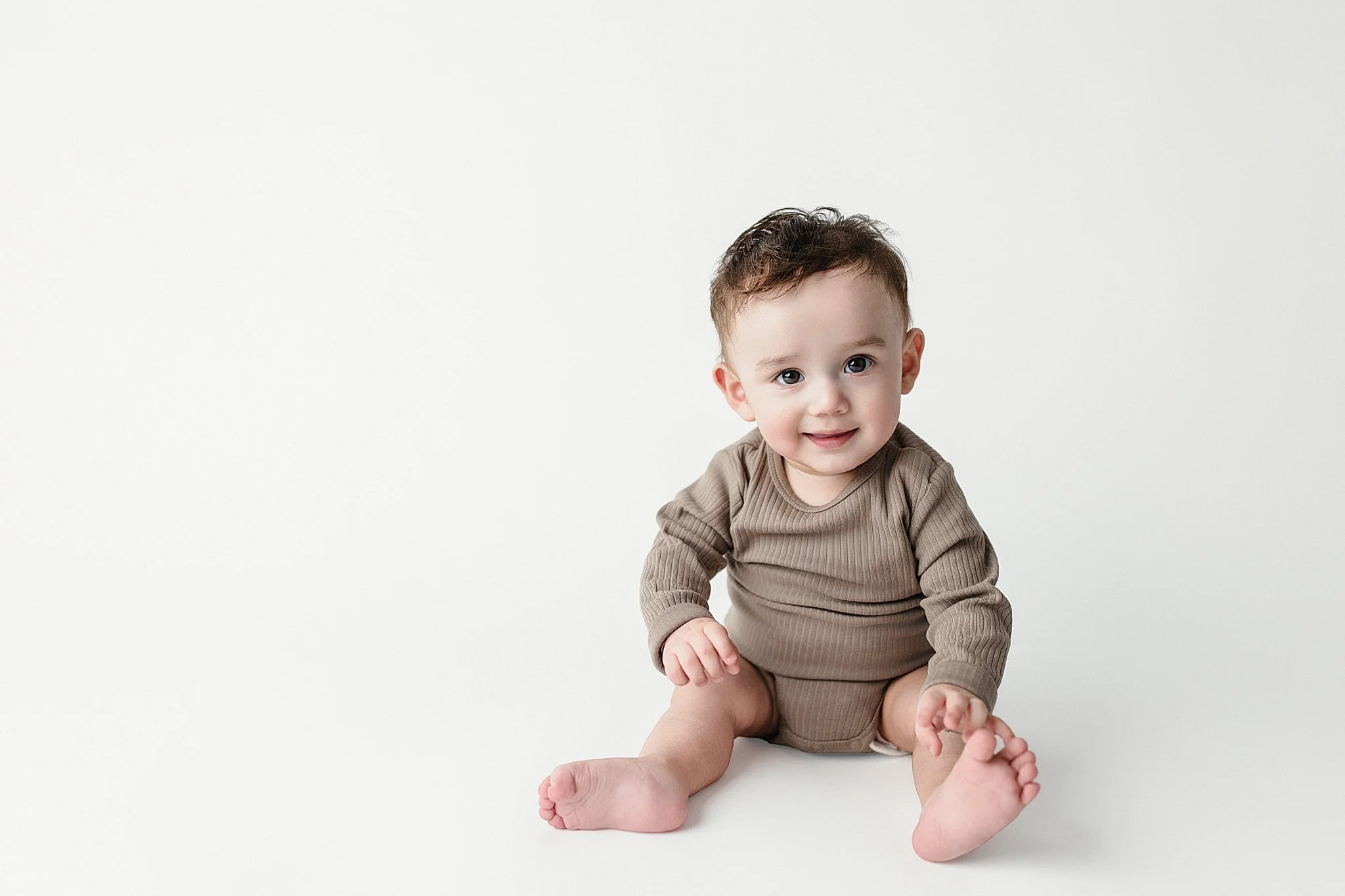 Cute baby sitting on a white background, wearing a beige long sleeve outfit, smiling and looking at the camera.