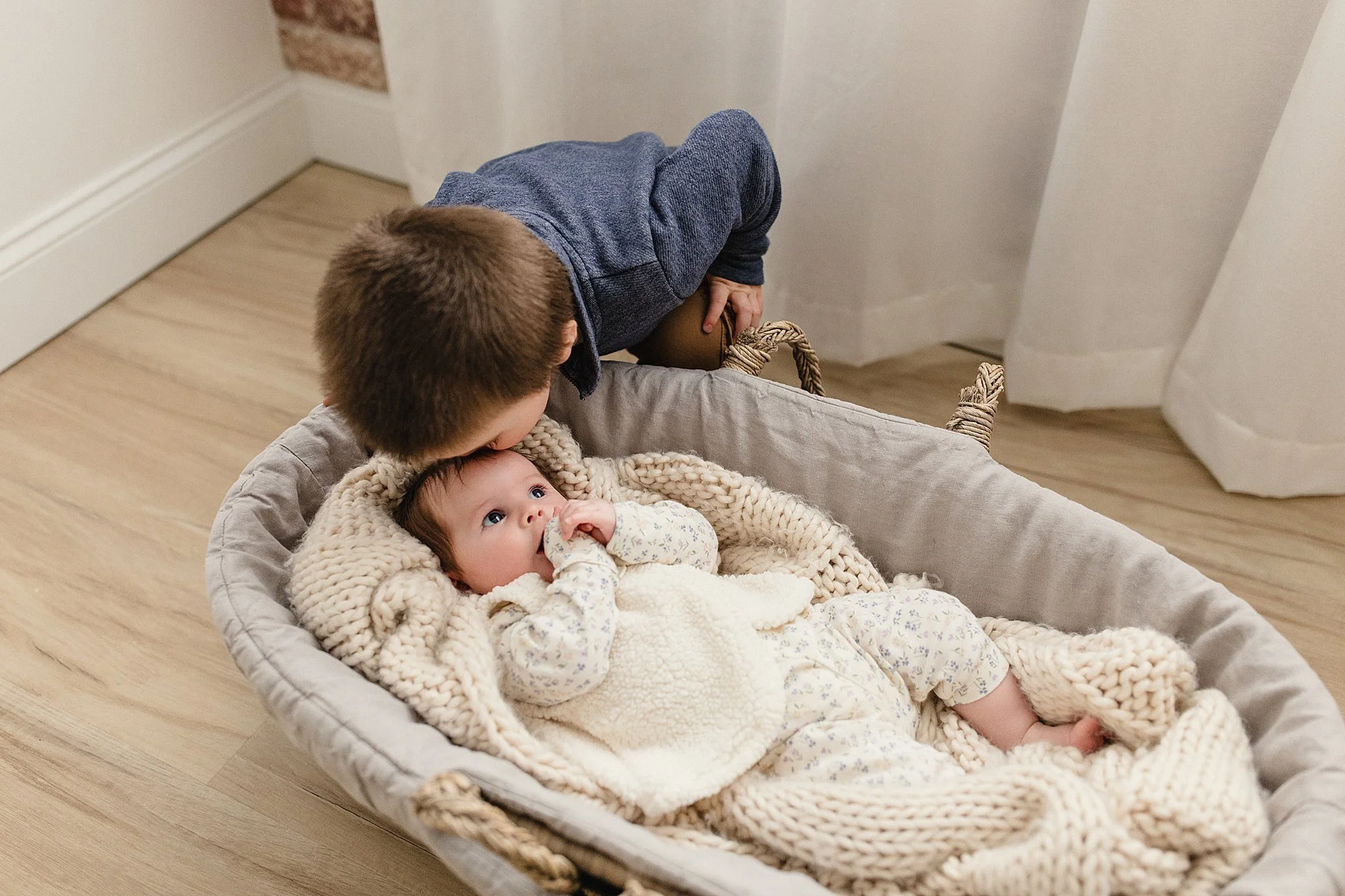 A young boy leaning over a baby girl lying in a gray woven basket, both indoors on a wooden floor, with the baby looking up at the boy, wrapped in cozy blankets during Naperville newborn photography.