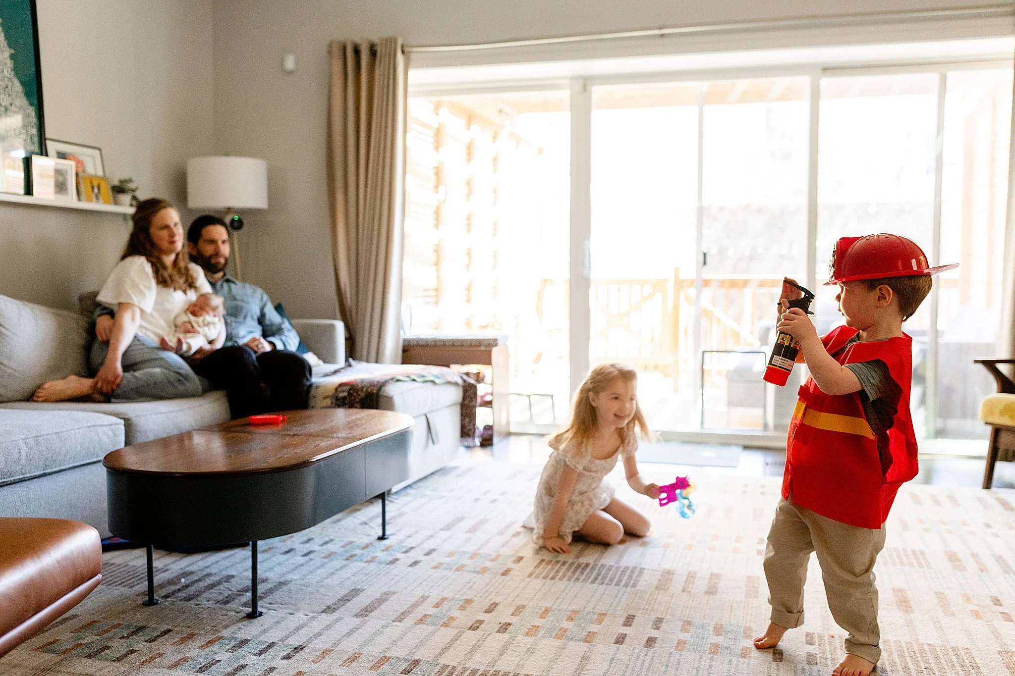 Family with two young children playing in living room, with parents sitting on sofa in background, bright sunlight coming through large sliding glass door during an in home family photo session in Naperville, IL.
