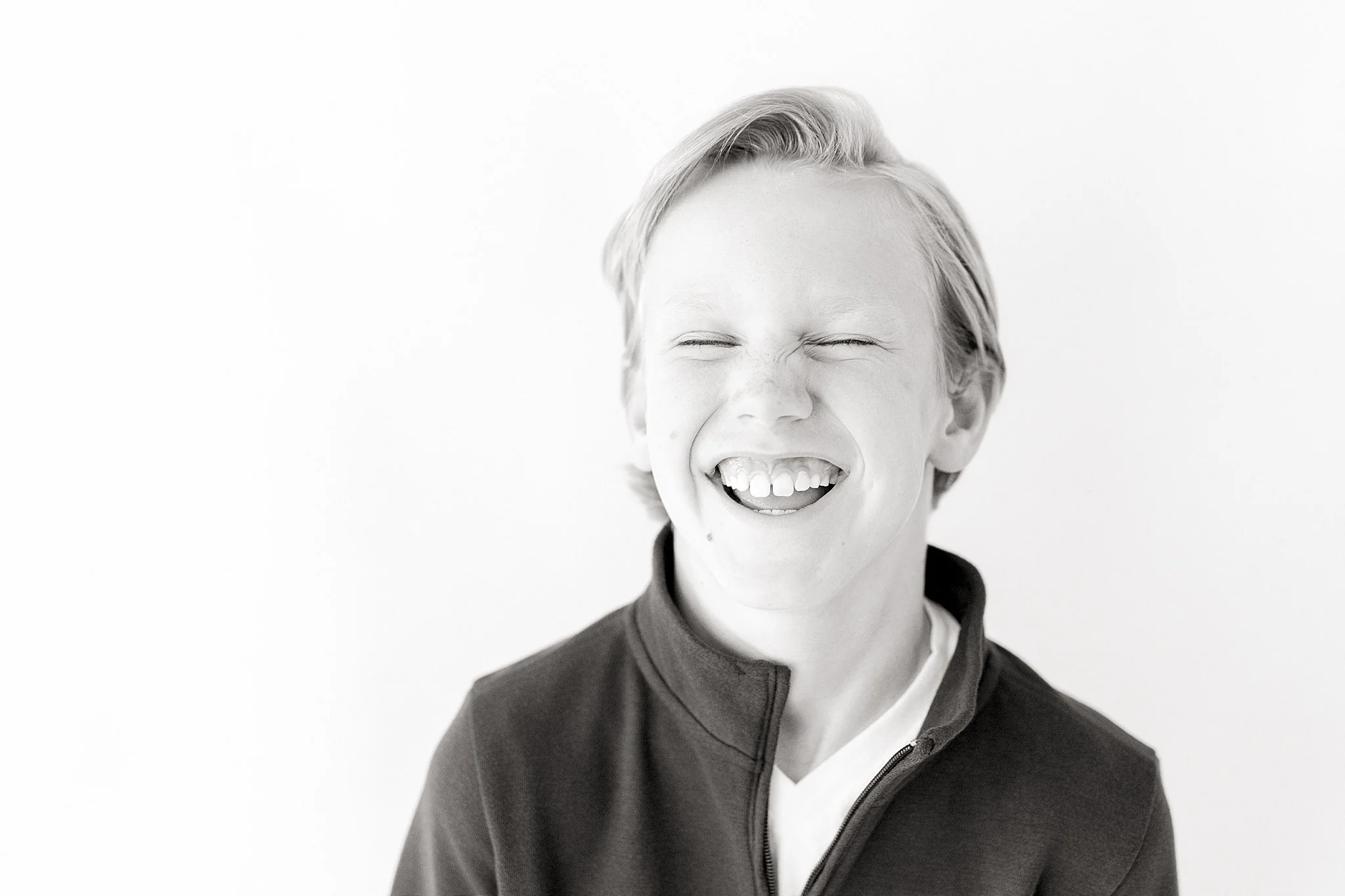 A young boy with blonde hair laughing with eyes closed, wearing a dark zip-up jacket and a white shirt, against a plain white background for kids acting photos in Naperville, IL.
