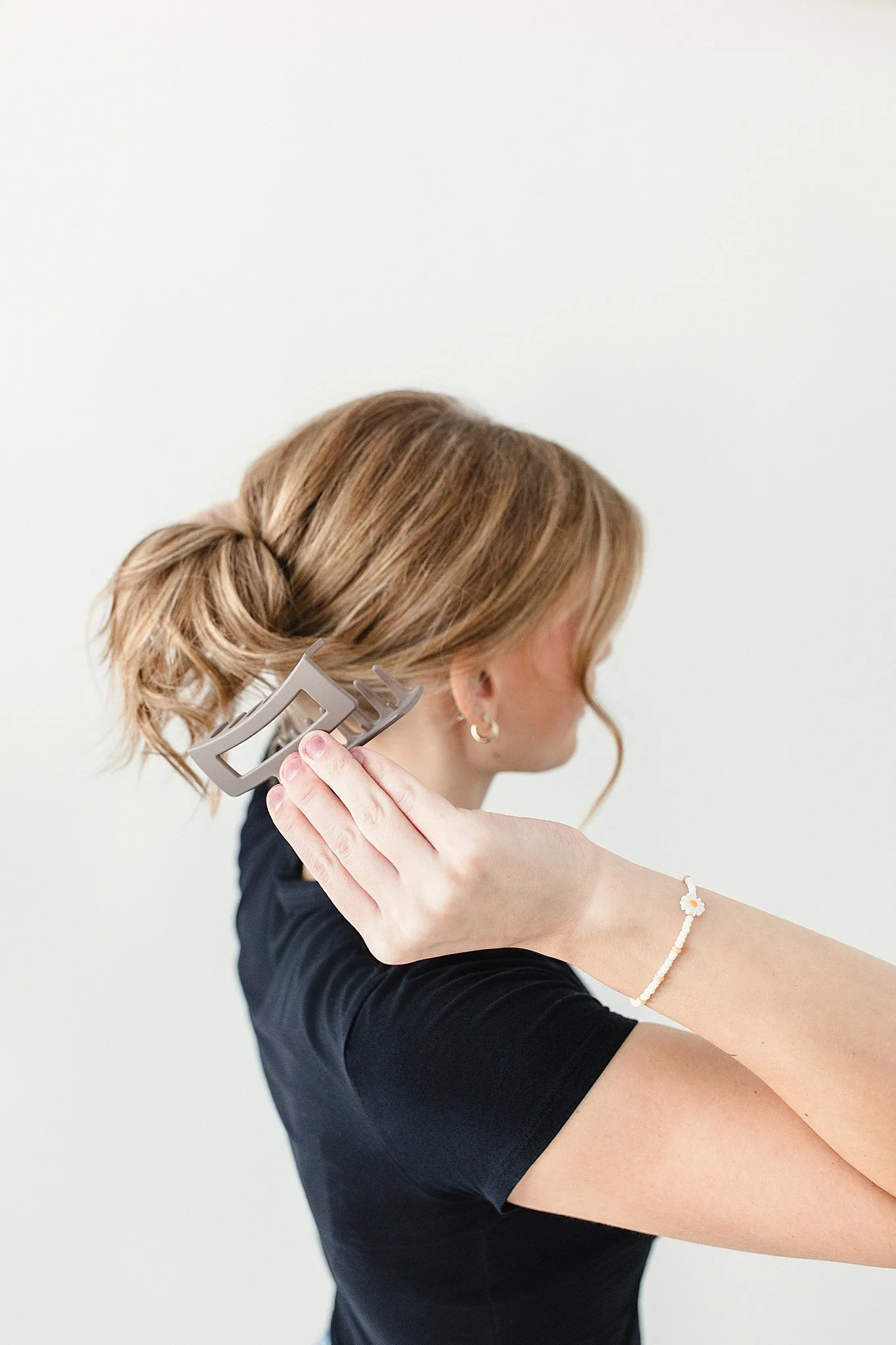 A woman with light brown hair styled in a loose bun is holding a hair clip in her hand and appears to be adjusting her hair. She is wearing a black top, earrings, and a white bracelet with a flower charm for hairstylist branding photos.