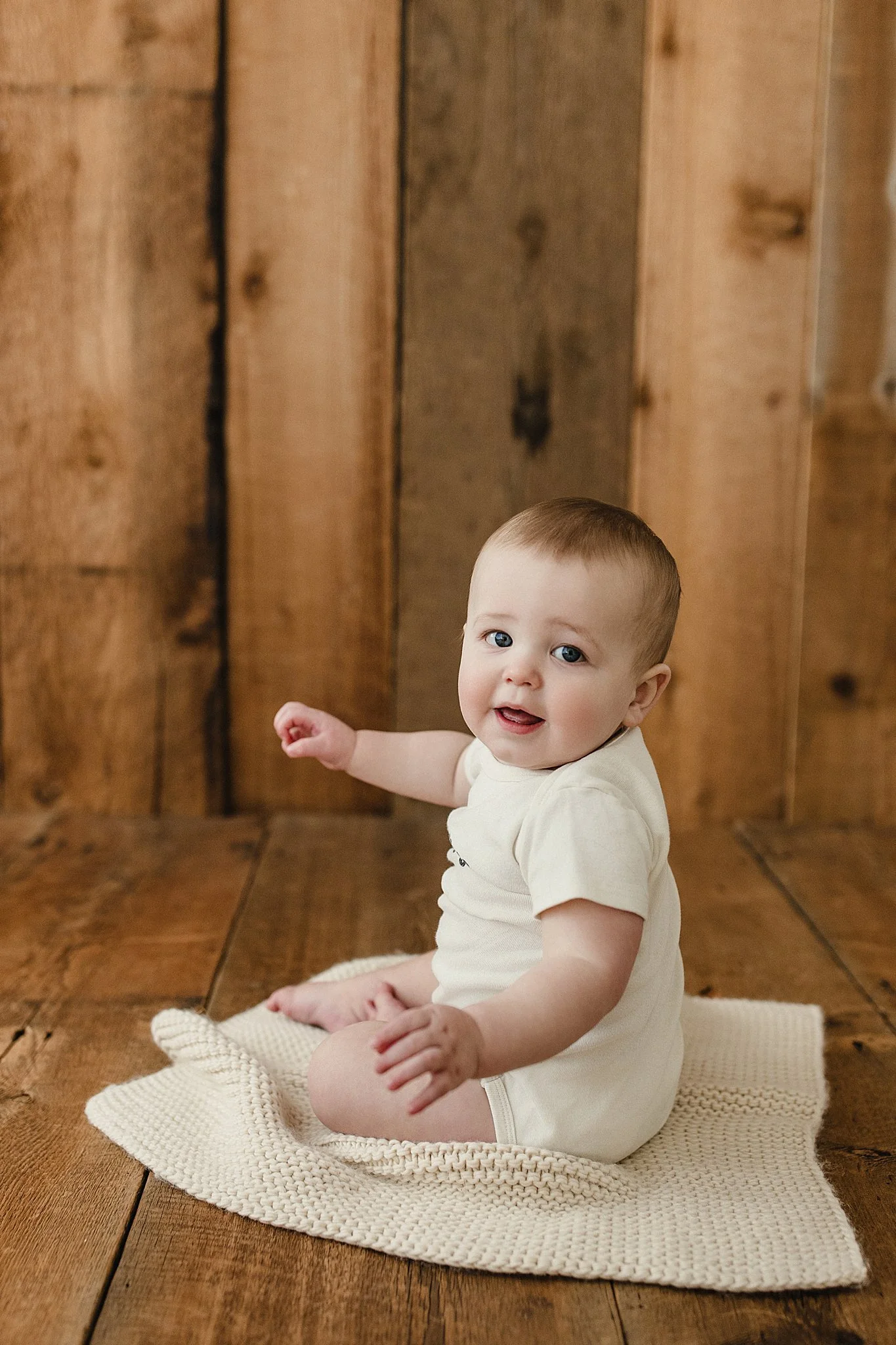 A baby sitting on a cream-colored knitted blanket on a wooden floor, with a wooden plank wall in the background during a milestone photoshoot for a first birthday in Naperville.
