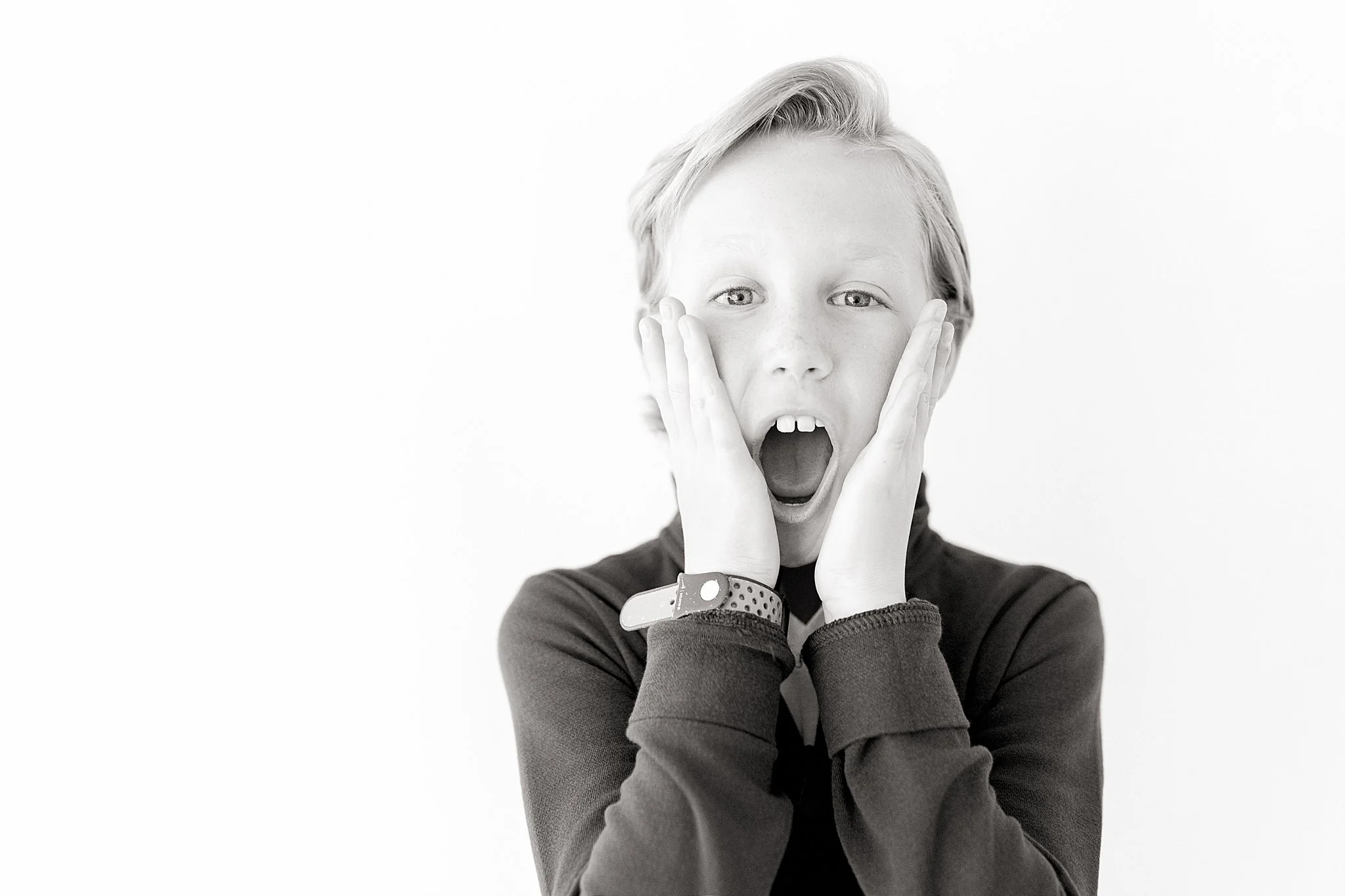 Black-and-white photo of a young person with short, light hair, mouth open in shock or surprise, hands on cheeks, wearing a dark sweatshirt and a wristband during personality minis with Ally and B Photography.