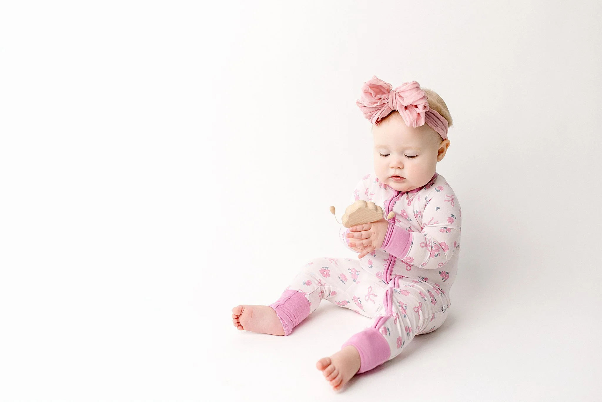 A young baby girl with a pink headband, sitting on a white surface, wearing pink and white pajamas with a bow pattern, holding a wooden toy with a small stuffed animal attached for first birthday photos near me.