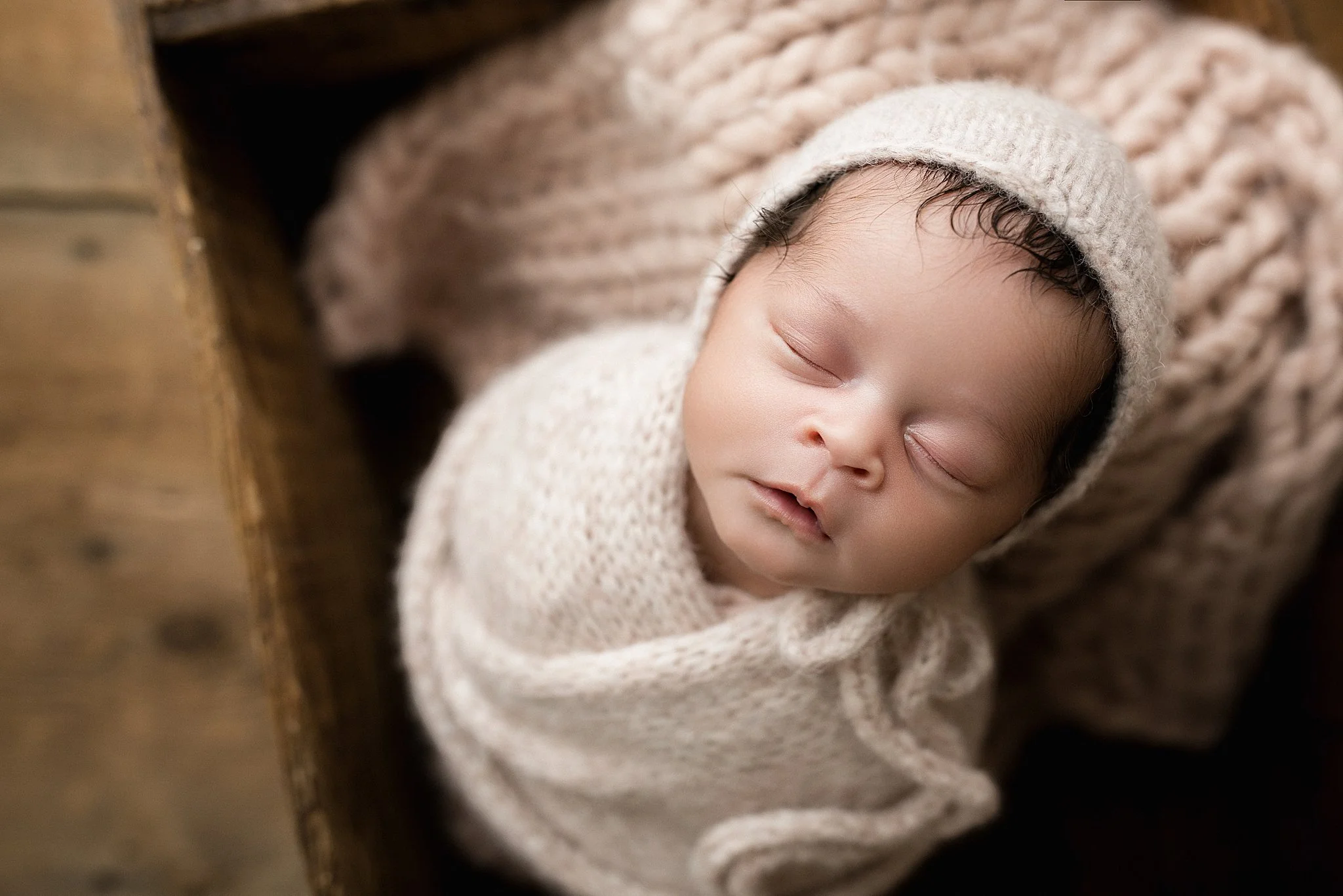 A sleeping infant with closed eyes, wearing a knitted hat, lying on a blanket with a textured pattern, with a wooden surface in the background at a photography studio in downtown Naperville, IL.