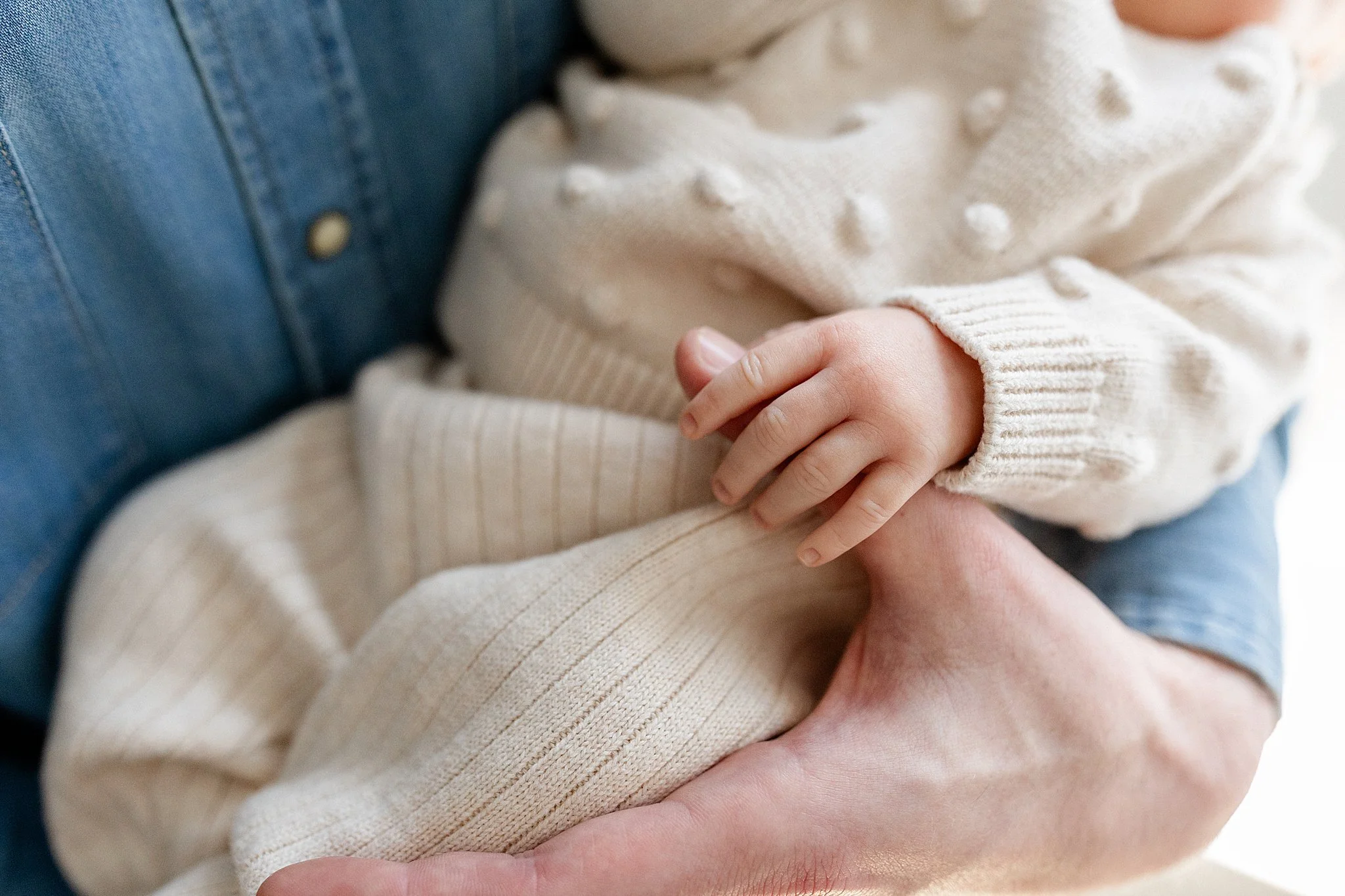 Close-up of a child's hand resting on an adult's arm, both wearing cozy, light-colored knit sweaters. The child is dressed in a cream-colored sweater with a textured pattern during lifestyle newborn photos Naperville.