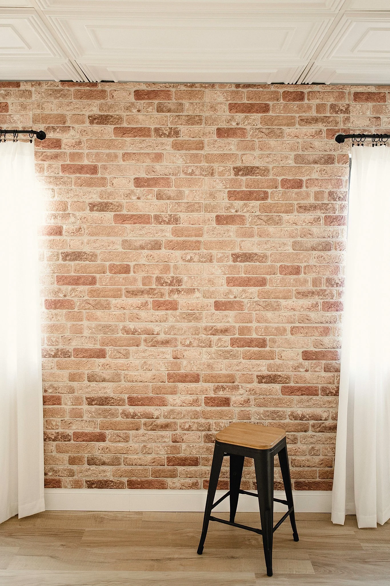 Empty room with a brick wall, white curtains on each side, and a wooden stool in the center in photography studios near me.