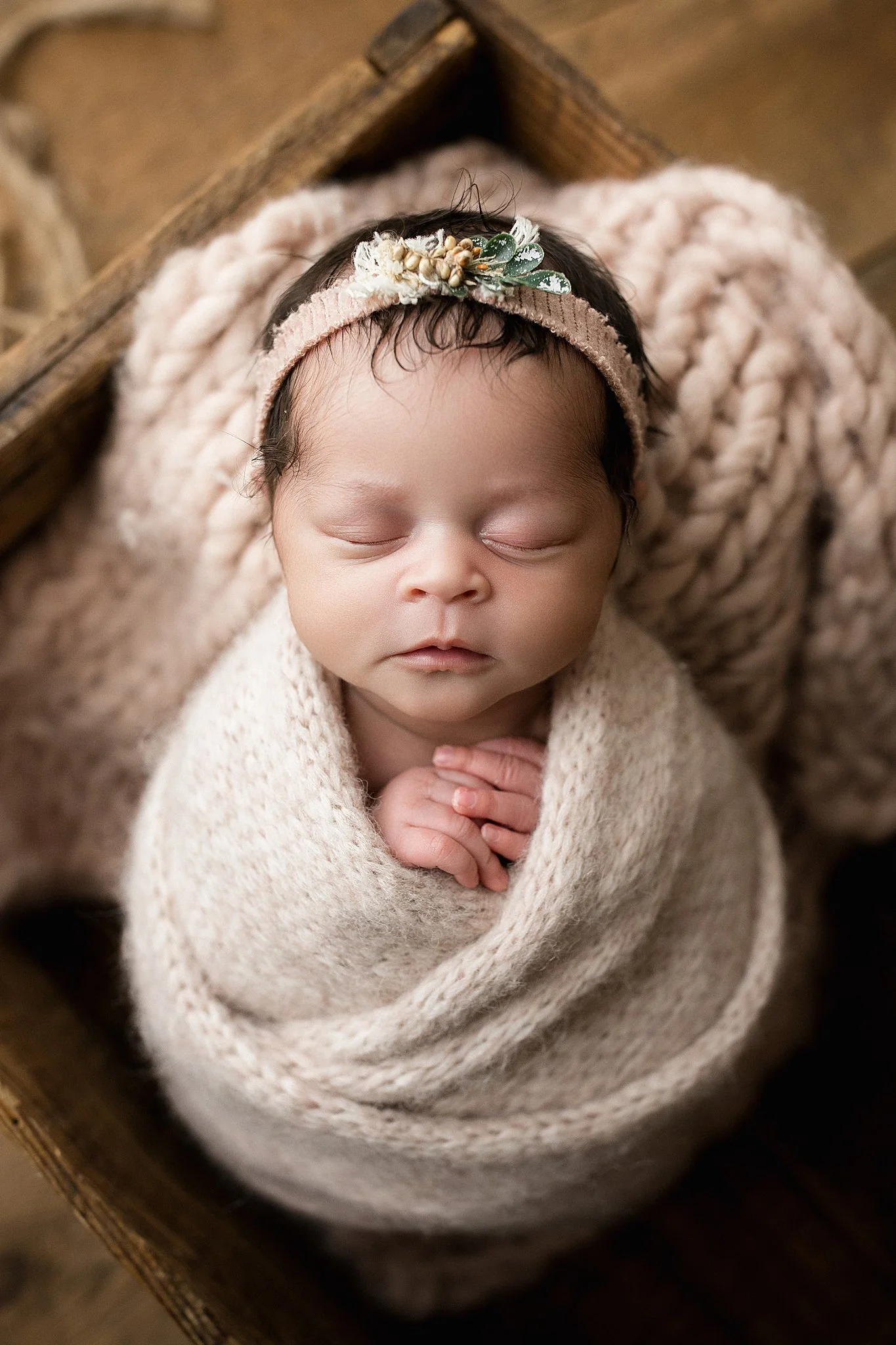 A sleeping newborn baby wrapped in a beige knit blanket, lying in a wooden basket, with a headband decorated with small flowers and greenery during newborn photography near me.
