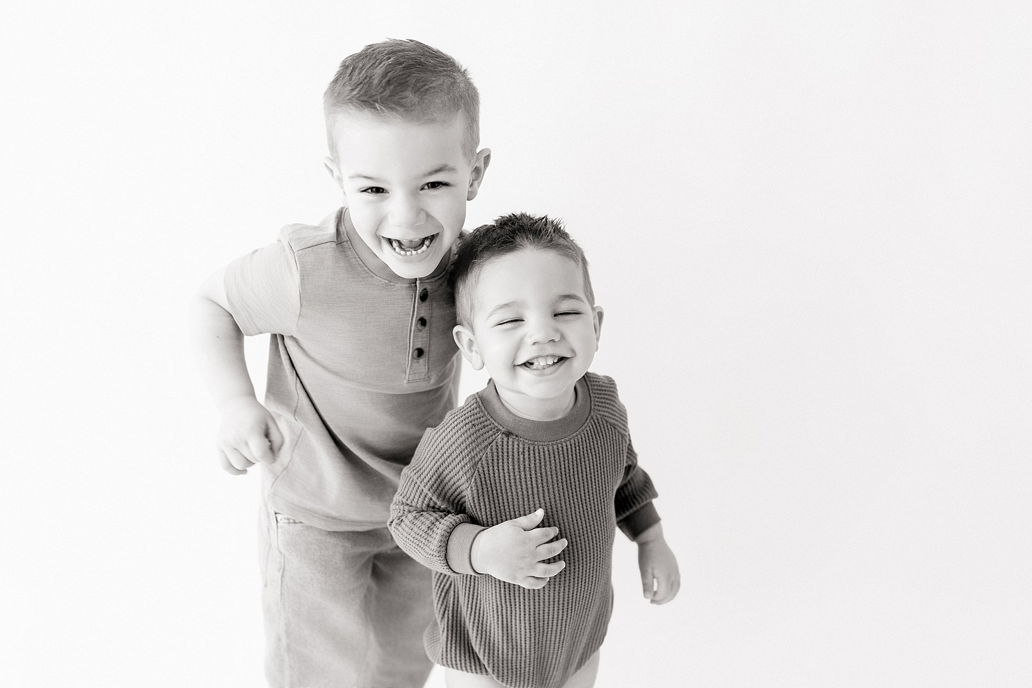 Two young boys smiling and playing together, one slightly older with short hair and the other younger. They are indoors with a plain background with a Naperville kids black and white portrait photographer.