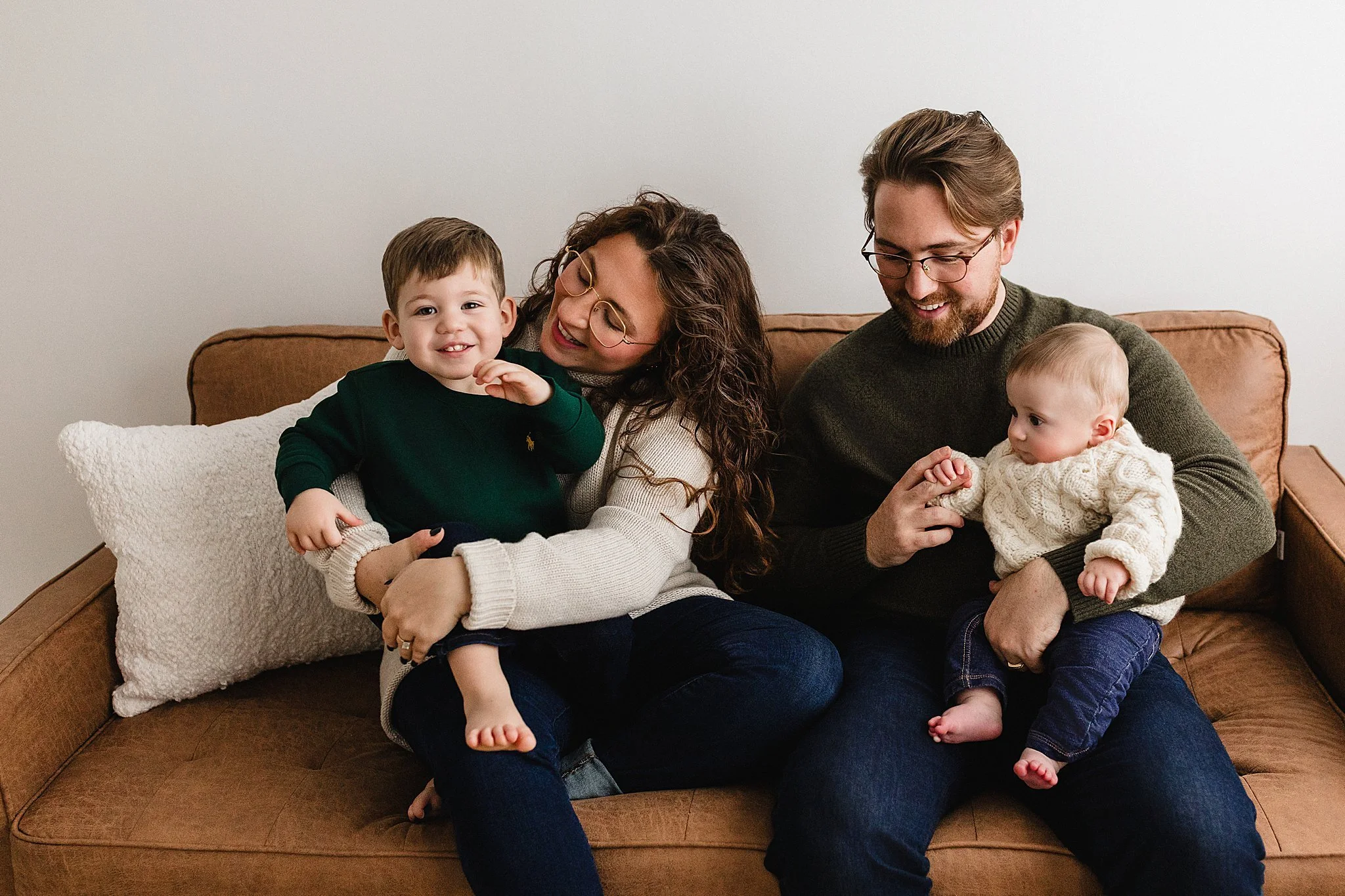 A family of four sitting on a brown couch, with the mother holding a young boy and the father holding a baby girl, all smiling and enjoying time together indoors for a Naperville family photography session near Naperville, IL.