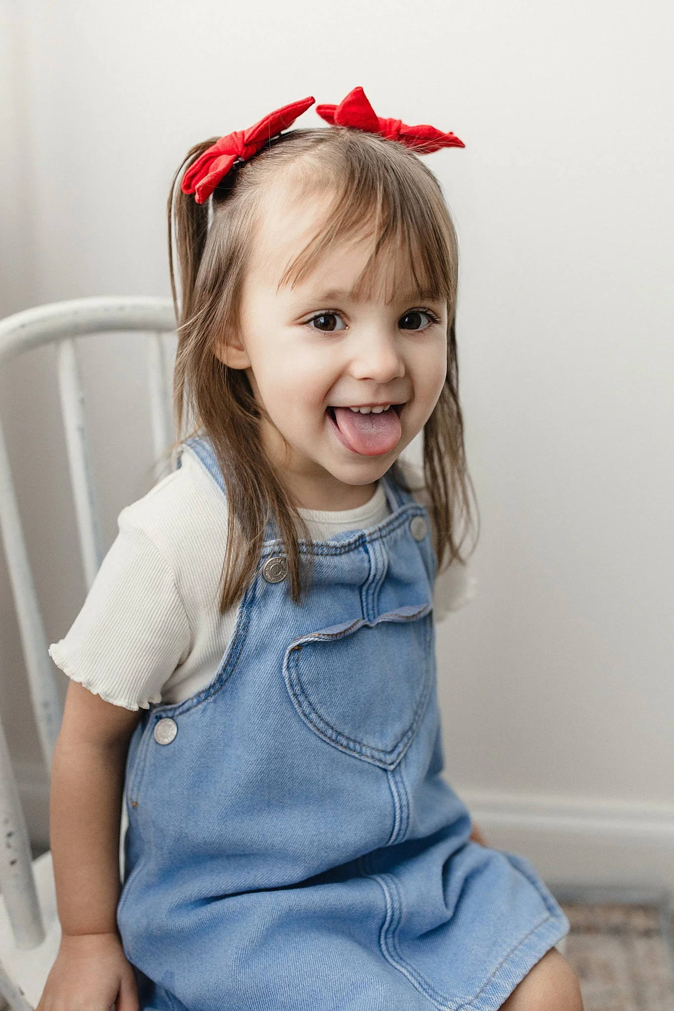 A young girl with brown hair and wearing a red bow, sticking out her tongue, sitting on a white chair with the best kids photographer in Naperville, IL.