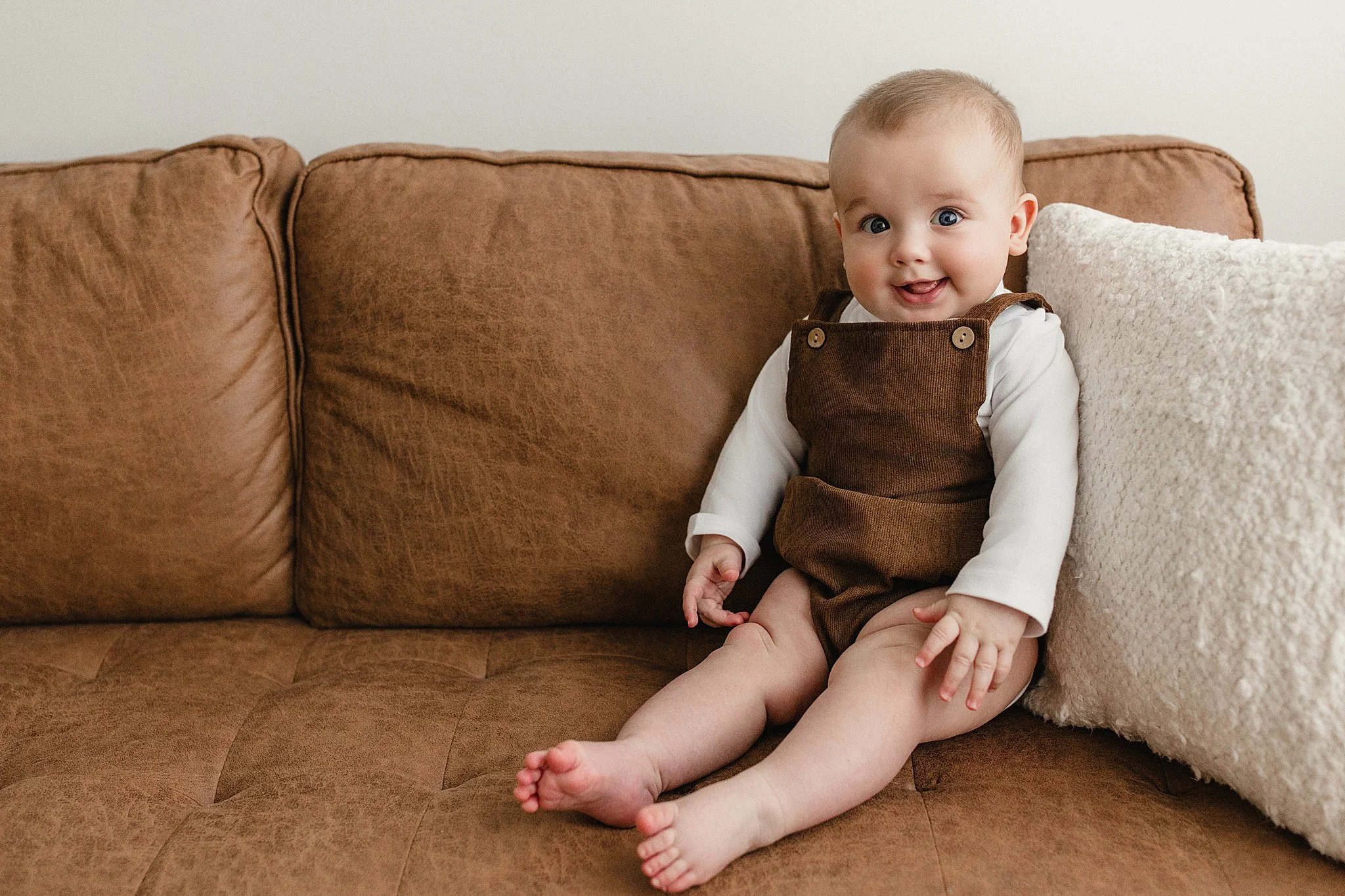 A happy baby girl sitting on a brown couch next to a cream pillow, smiling and looking at the camera during baby photoshoot with Naperville, IL.
