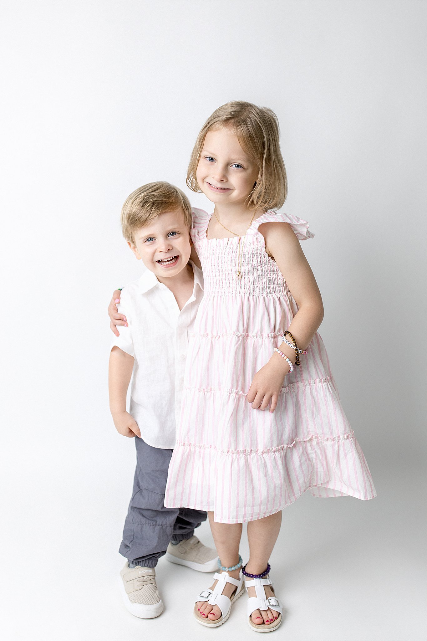 A young girl with shoulder-length blonde hair and a young boy with short blonde hair smiling while standing against a plain white background. The girl is wearing a pink and white striped sundress with jewelry, and the boy is dressed in a white shirt 