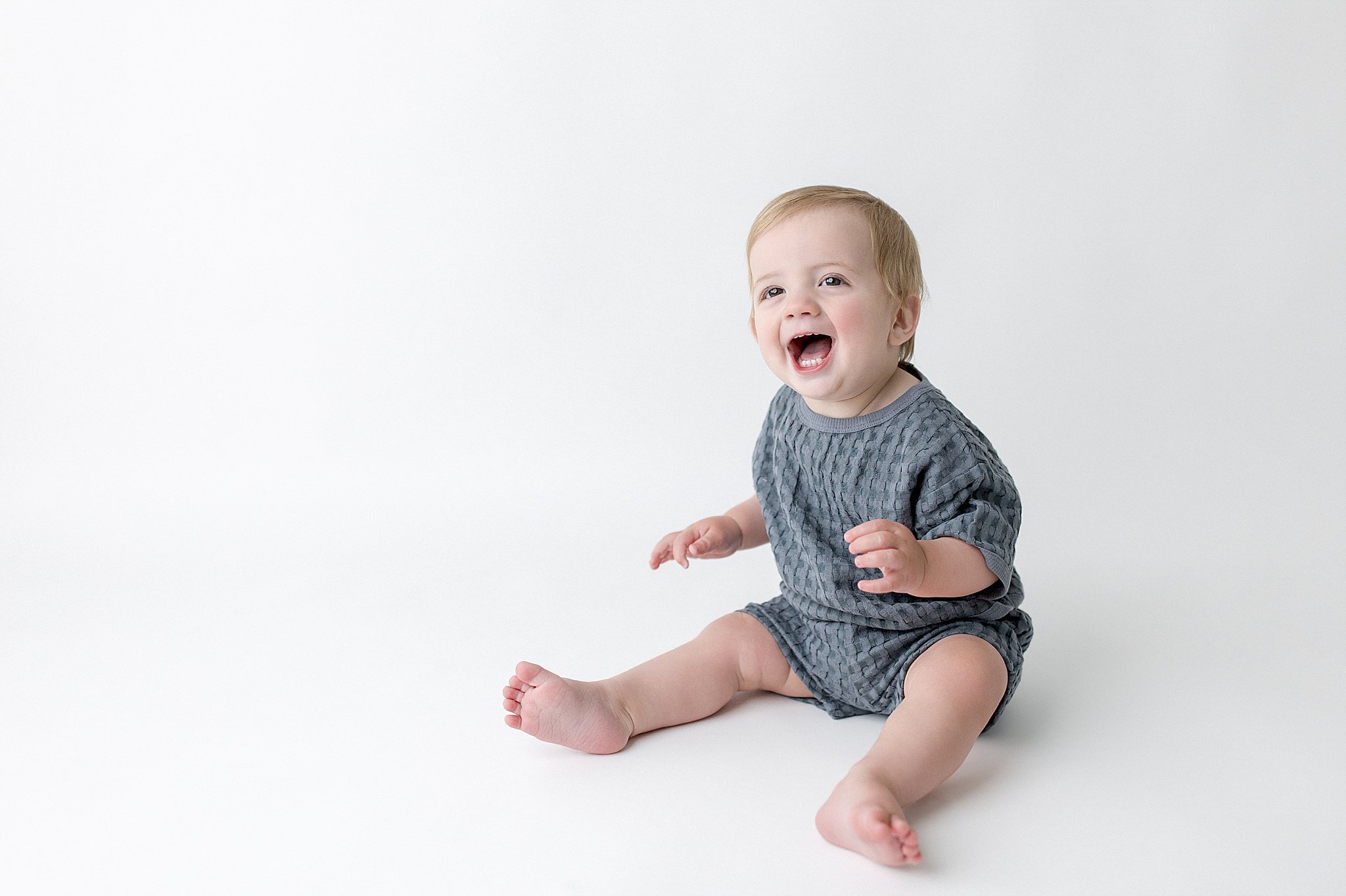 A smiling toddler with blonde hair, wearing a gray textured shirt and shorts, sitting on the floor against a plain white background during a milestone photography session in Naperville.