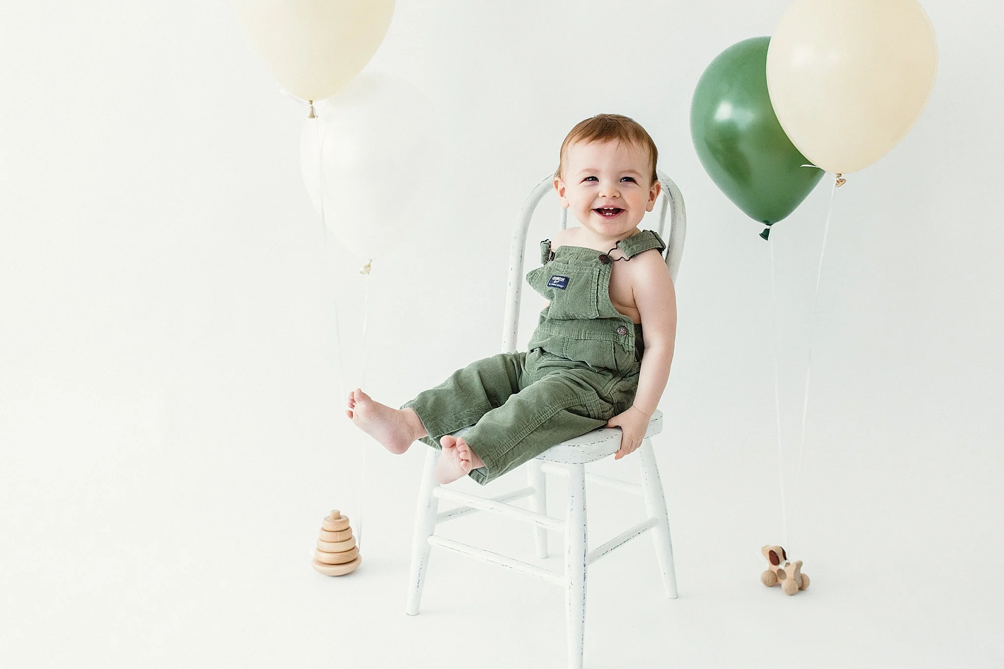 A smiling baby sitting on a white wooden chair surrounded by beige and green balloons, with a wooden toy on the floor for milestone photos near me.