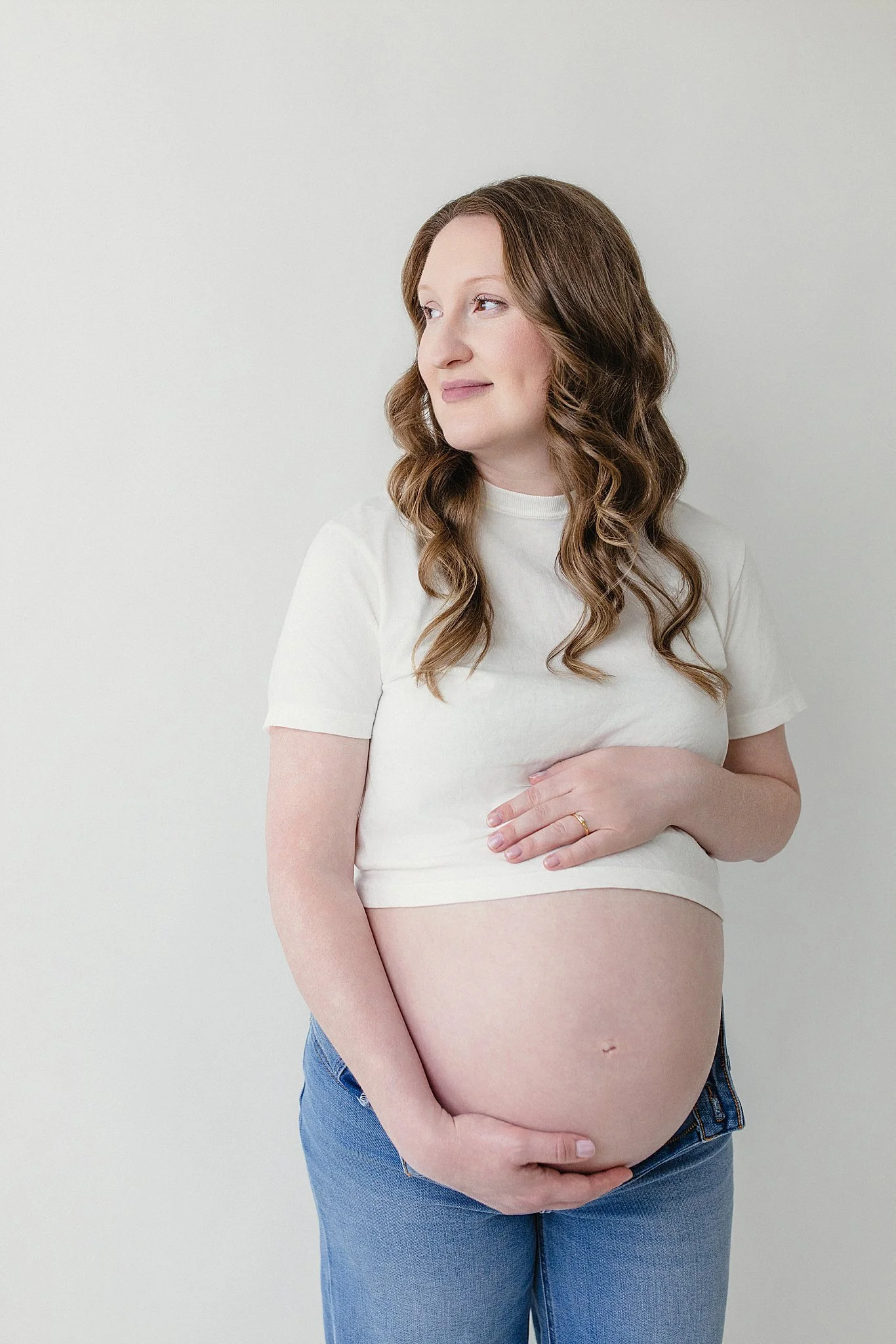 A pregnant woman with long wavy brown hair, wearing a light-colored t-shirt and blue jeans, cradling her belly with both hands, standing against a plain white wall with a Naperville maternity photographer.