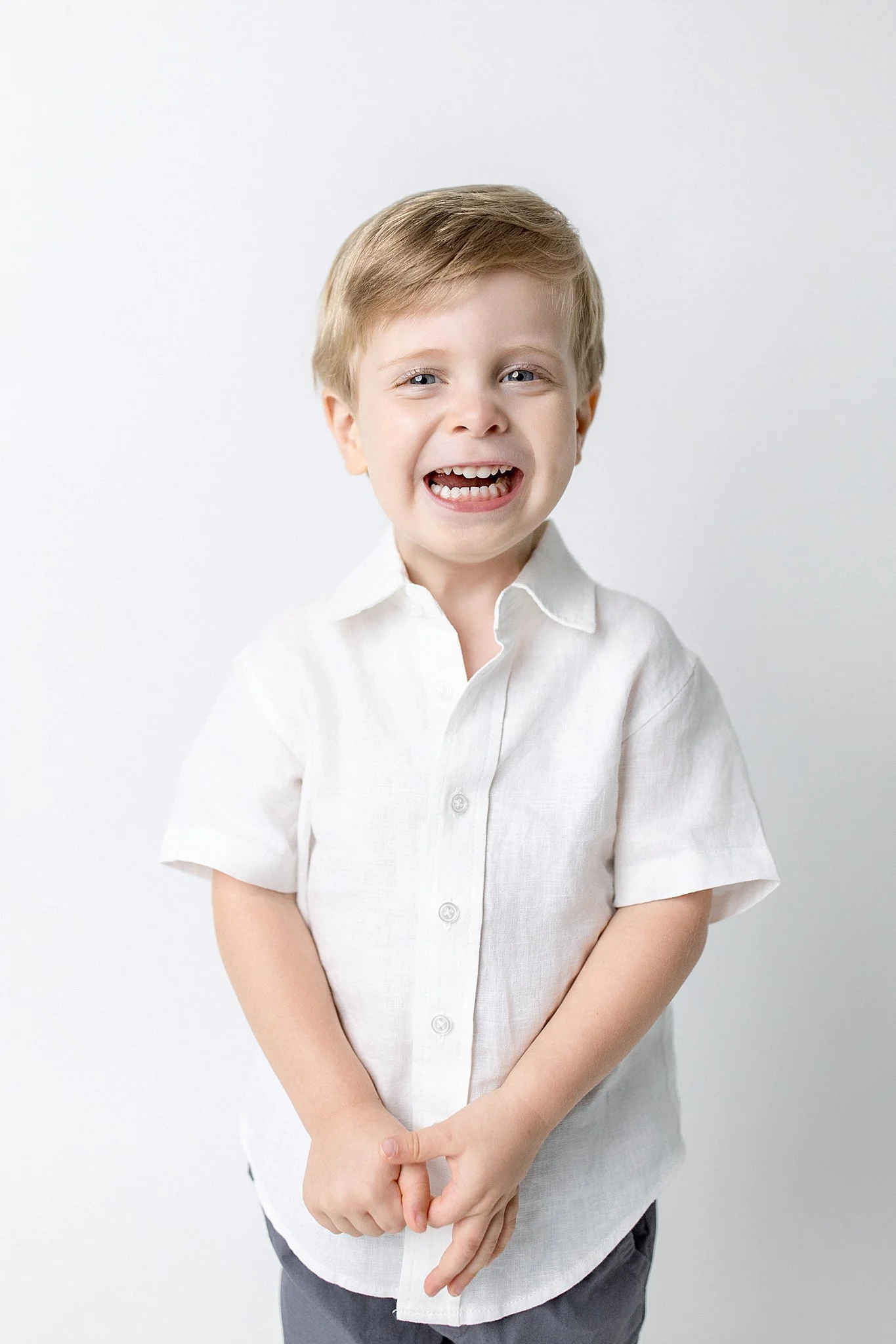 A young boy with blonde hair smiling and laughing, wearing a white button-up shirt and gray pants, standing against a plain white background with a personality mini session photographer near me.