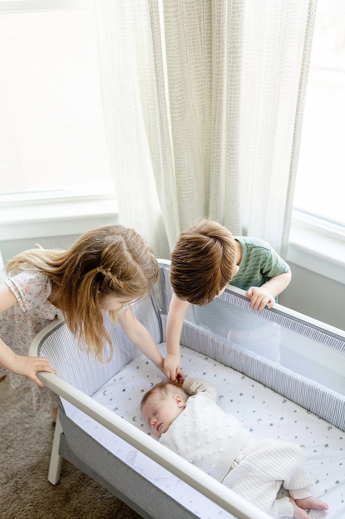 Four children, three girls and one boy, are gathered around a crib with a sleeping baby inside. The children are reaching into the crib, and they appear to be interacting and looking at the baby. The crib has a star-patterned sheet, and the room has 