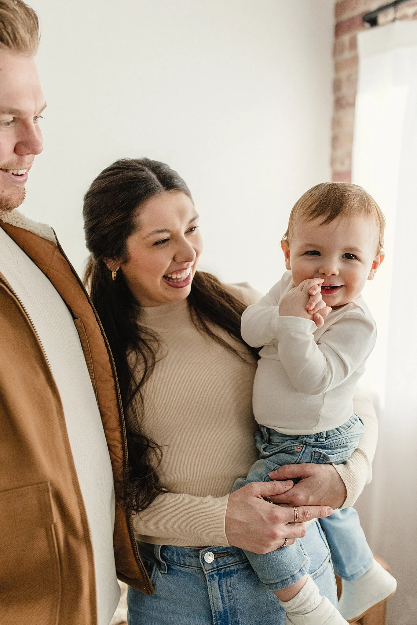 A family of three, including a woman holding a toddler, smiling and interacting with a man, in a cozy indoor setting during family lifestyle photos in Naperville, IL with Ally and B Photography.