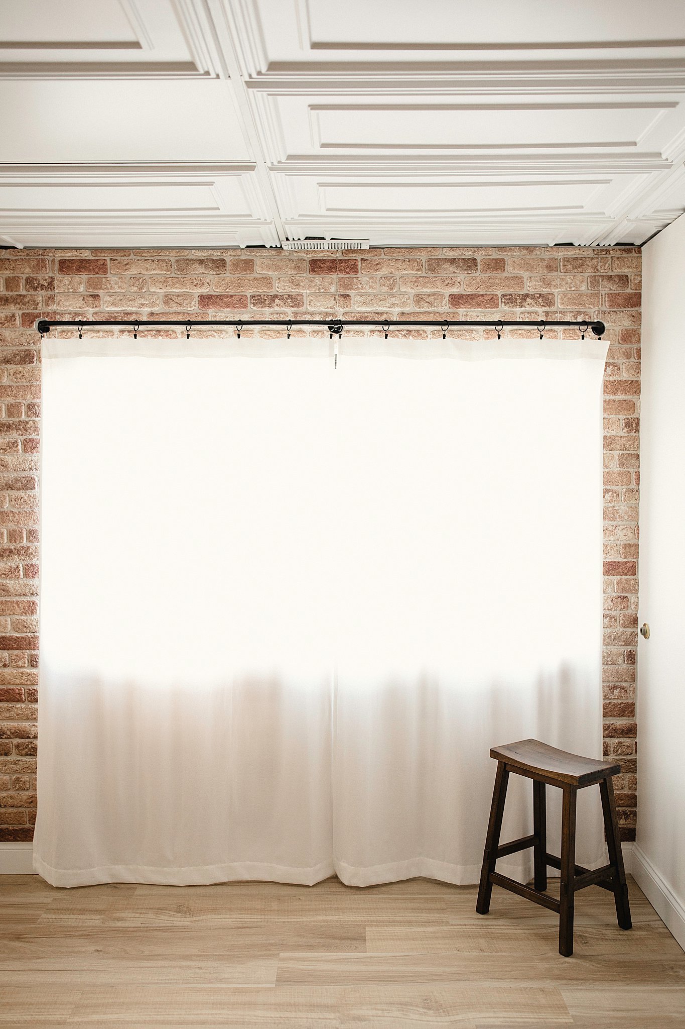 Interior shot of a room with a brick wall and white ceiling, featuring a white curtain hanging on a black rod, and a wooden stool on the floor in Ally and B Photography studio Naperville.