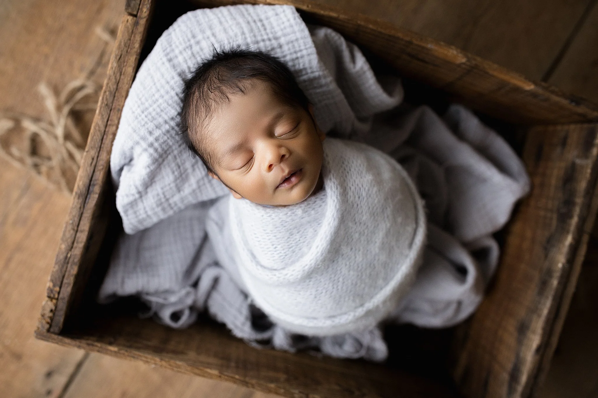 A sleeping baby wrapped in a white blanket, lying in a wooden crate, with a gray checkered cloth beneath their head during a posed newborn photo session.