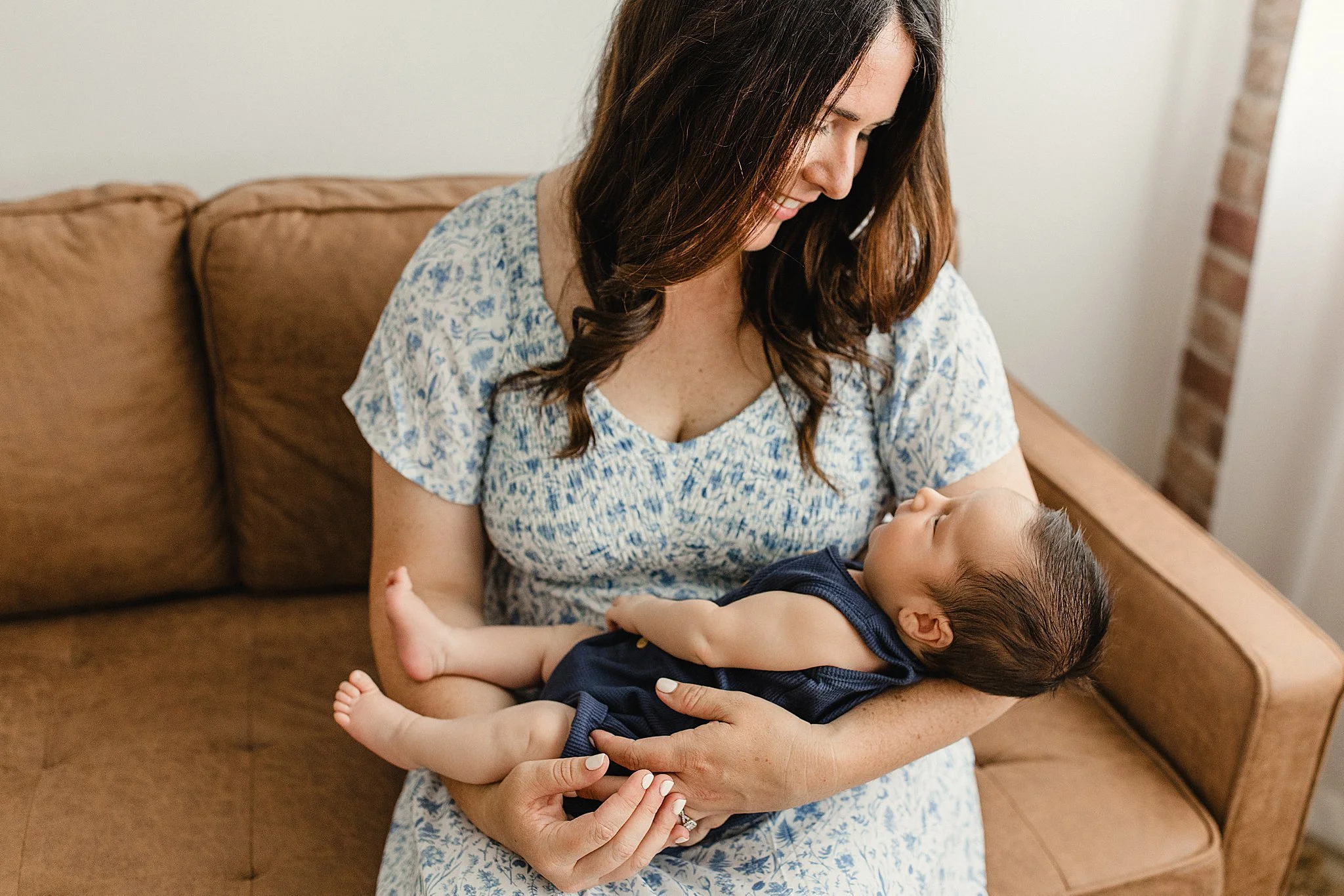 A woman with long brown hair smiling while holding a small child lying in her arms on a brown couch in a living room for lifestyle newborn photos with Ally and B Photography.