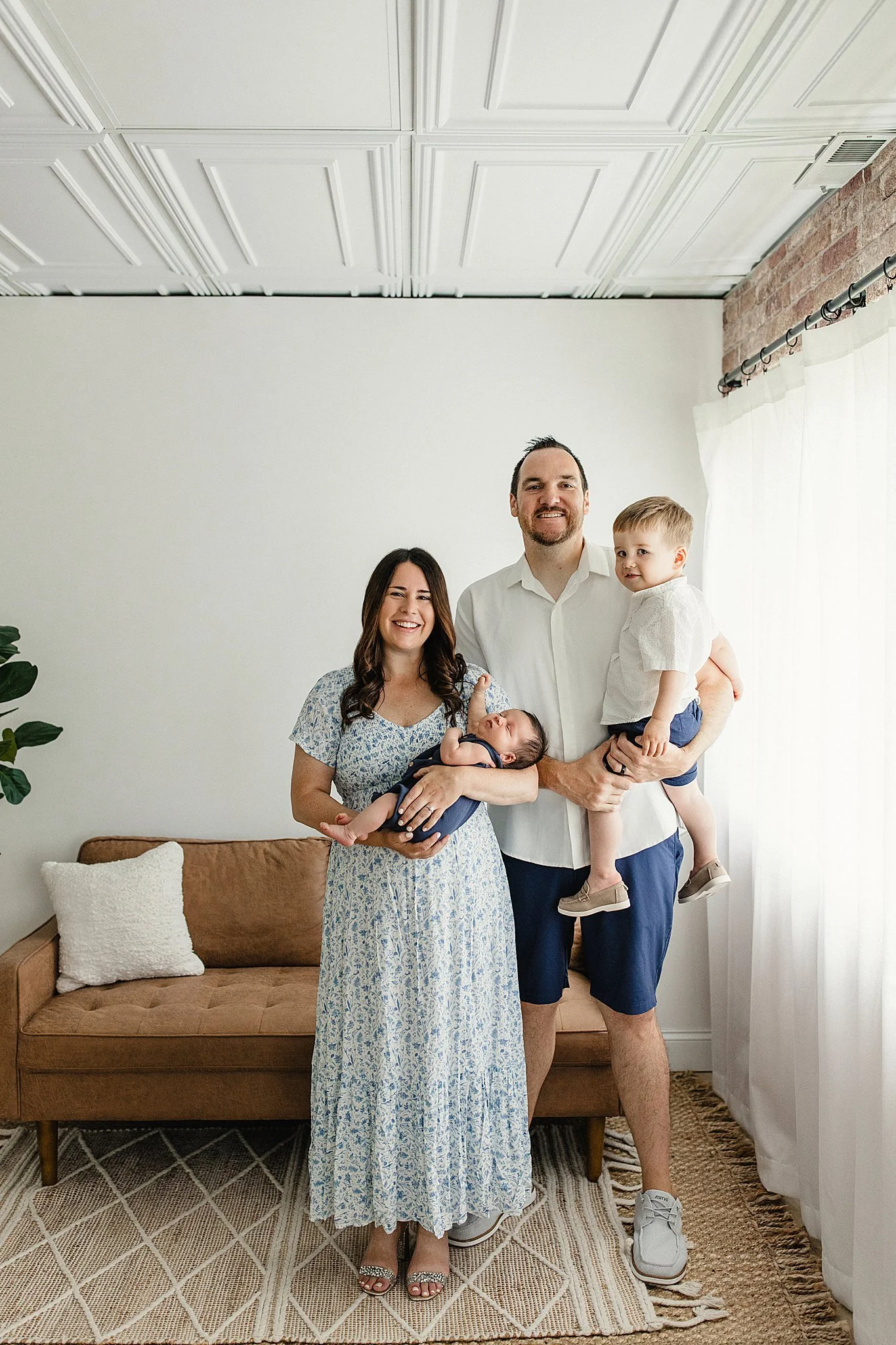 A family of four standing in a living room, with a woman in a long blue patterned dress, a man in a white shirt and navy shorts holding a young boy, and a newborn baby in the woman's arms. The family is smiling during a lifestyle photography session.