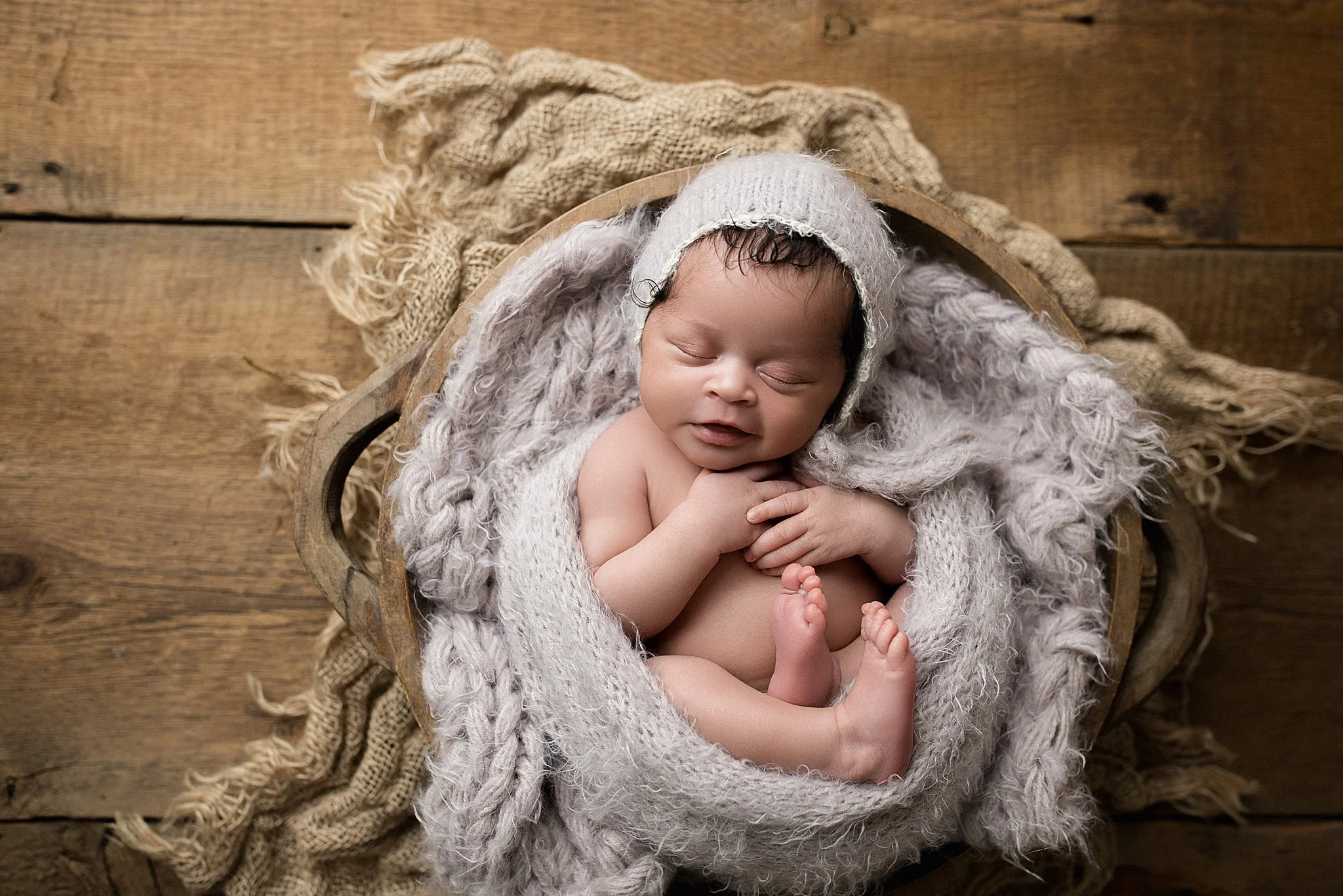 A sleeping baby with a knitted bonnet, wrapped in a blanket, lying in a wooden bowl on a rustic wooden surface with a beige cloth during a newborn session with Naperville newborn photography.