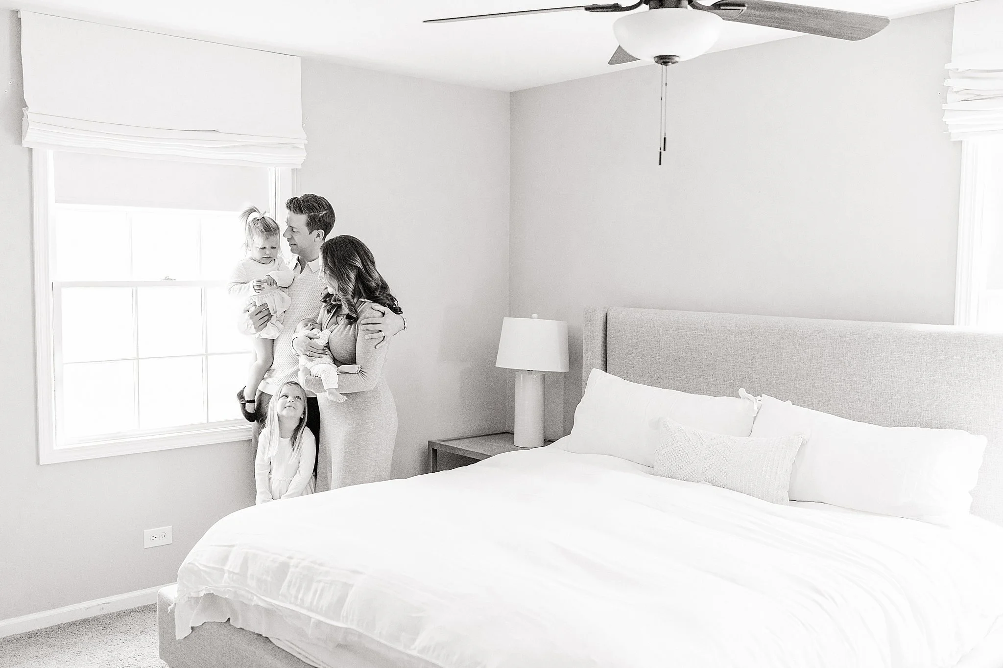 A black-and-white photo of a family of four standing by a bedroom window. The parents are each holding two young children, while the older daughter stands beside them looking up during a Naperville in home lifestyle newborn photo session.