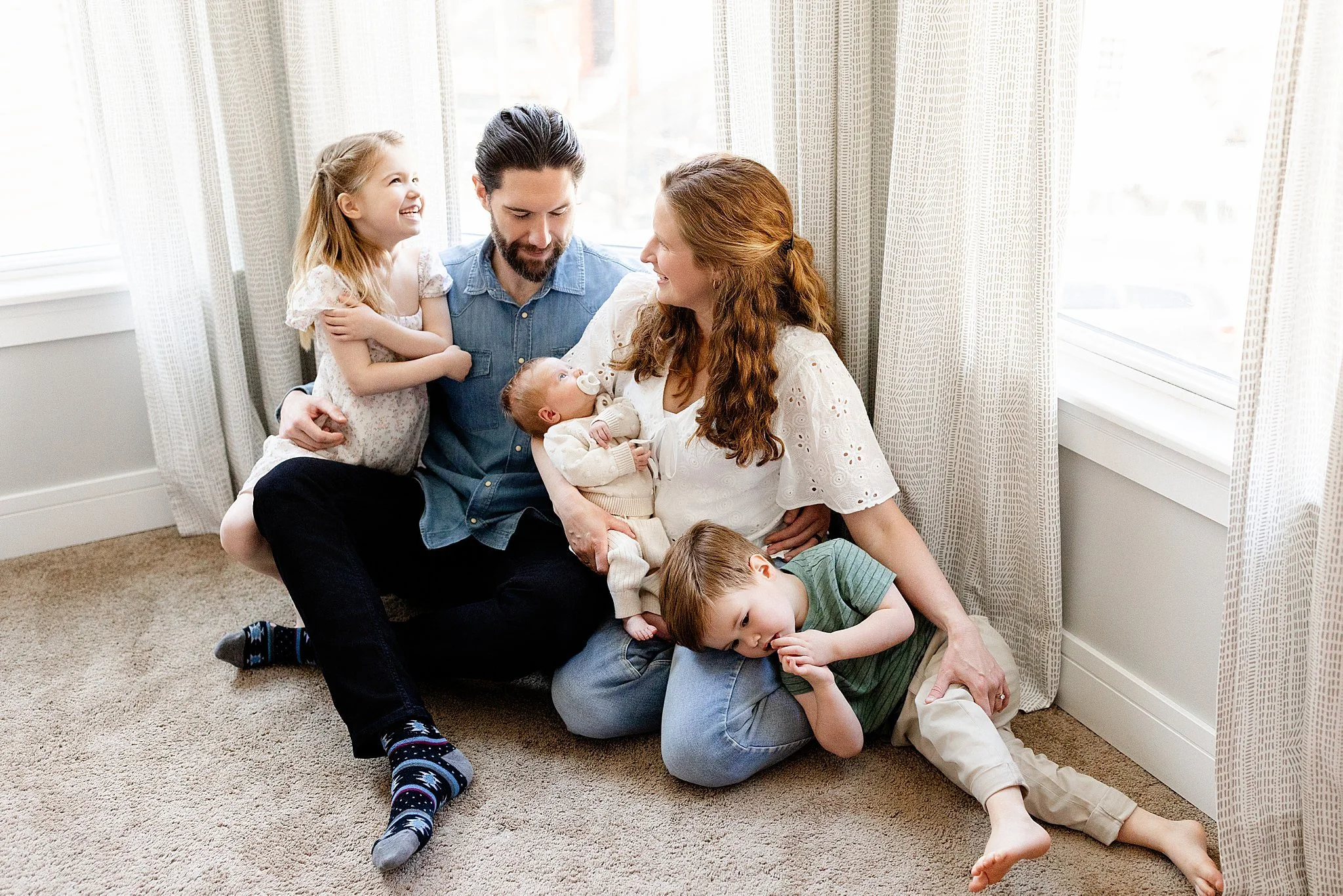 A family of six enjoying quality time together, sitting on a carpeted floor in front of a window with curtains. The parents are holding their infant, while the children are gathered around, smiling and interacting affectionately.