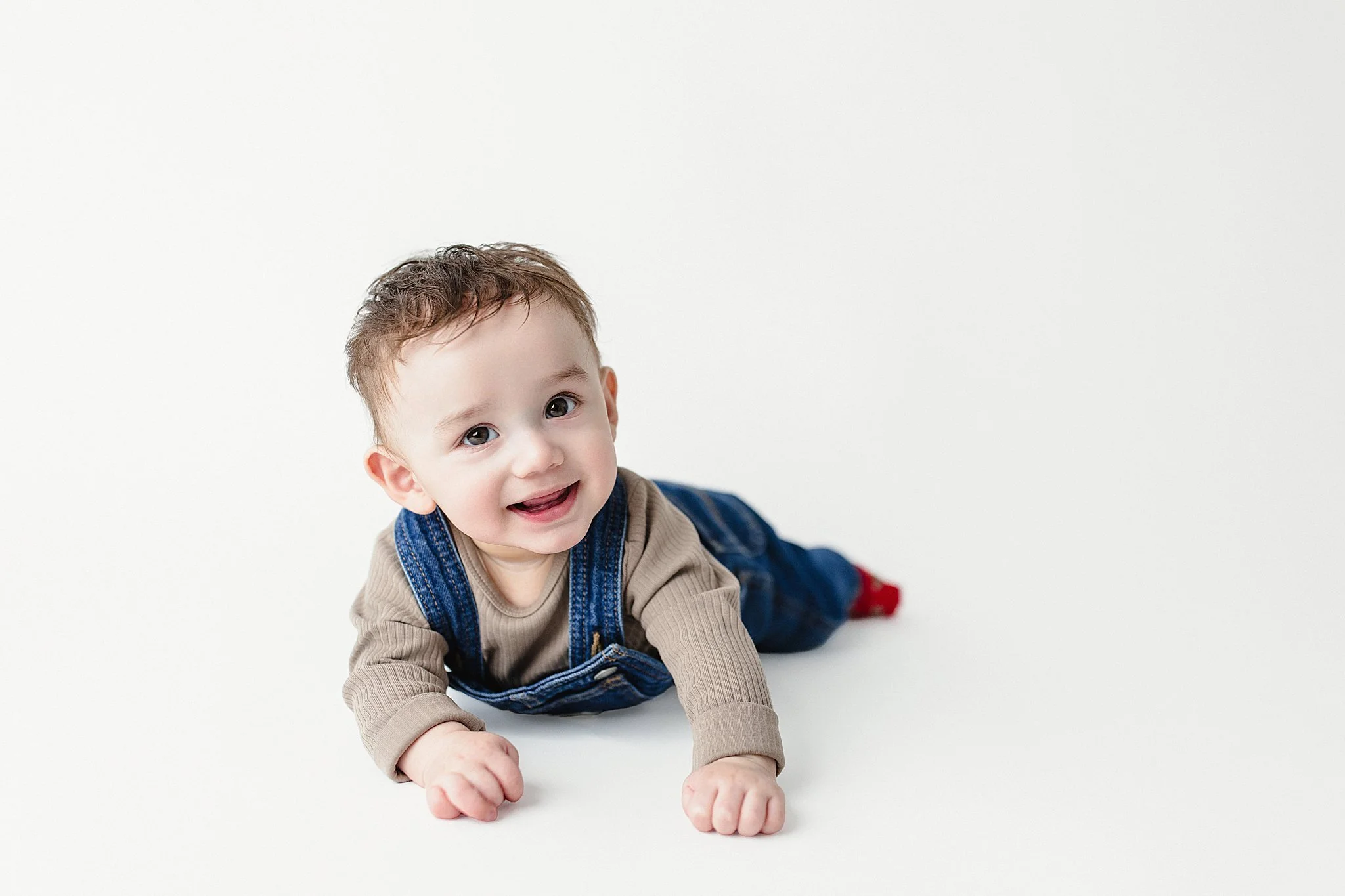 Smiling baby boy crawling on a white background, wearing a beige long sleeve shirt, denim overalls, and red socks during a mini milestone session with Ally and B Photography.