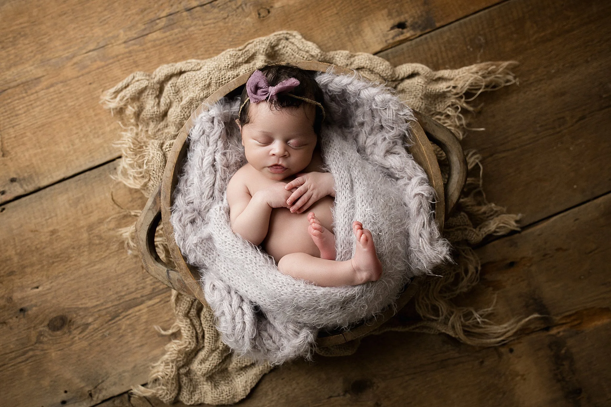 A sleeping baby with a pink bow on her head, wrapped in a fluffy blanket in a wooden basket, on a rustic wooden floor with a frayed burlap cloth underneath with newborn photography Naperville, IL.