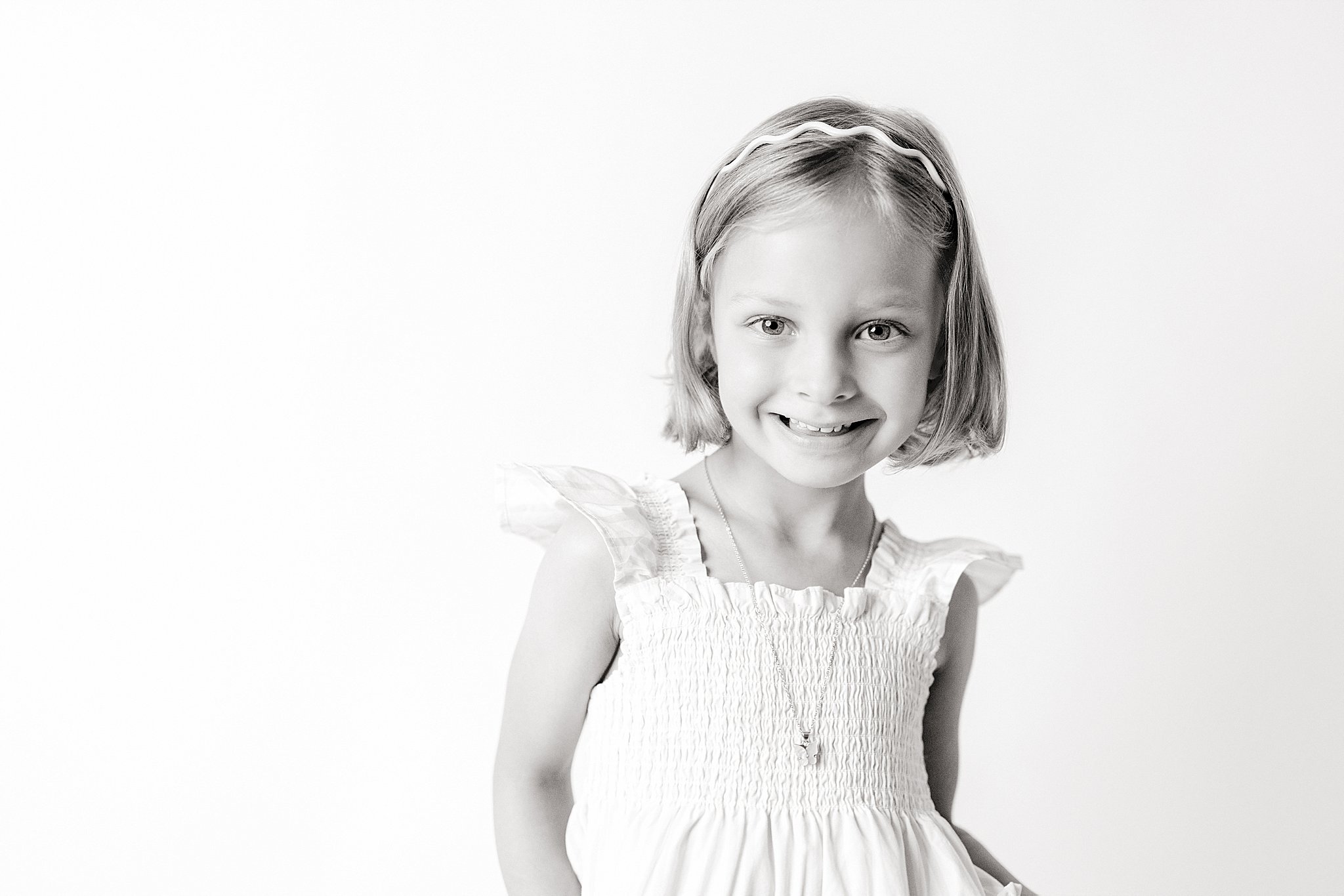 A smiling young girl with shoulder-length blonde hair, wearing a white dress with ruffled sleeves and a zipper detail, standing against a plain white background for black and white kids portraits near me.