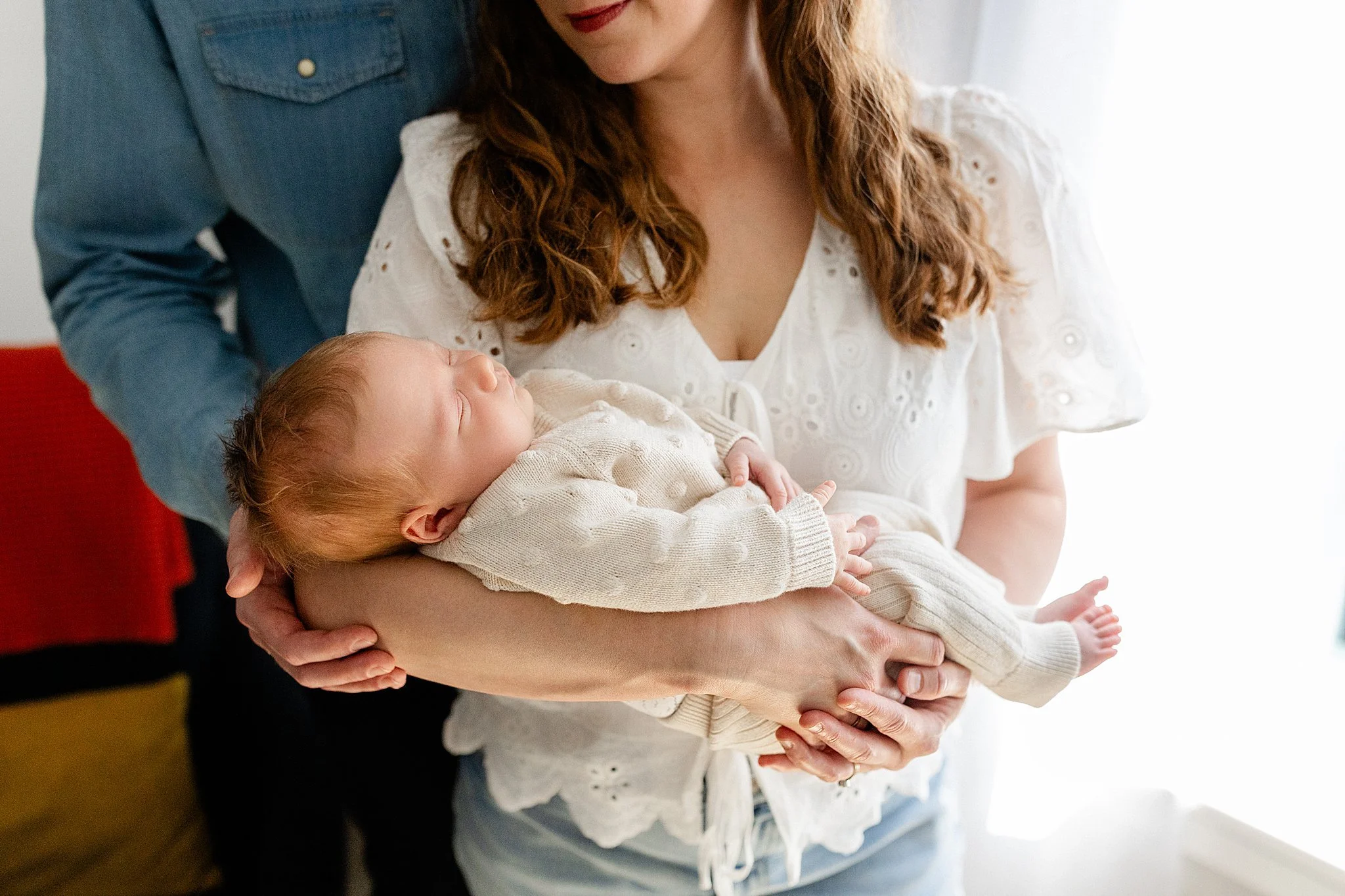 A woman holding a sleeping baby in her arms, with a man standing beside her. The woman is wearing a white blouse with eyelet details, and the man is wearing a denim shirt during lifestyle photos near me.