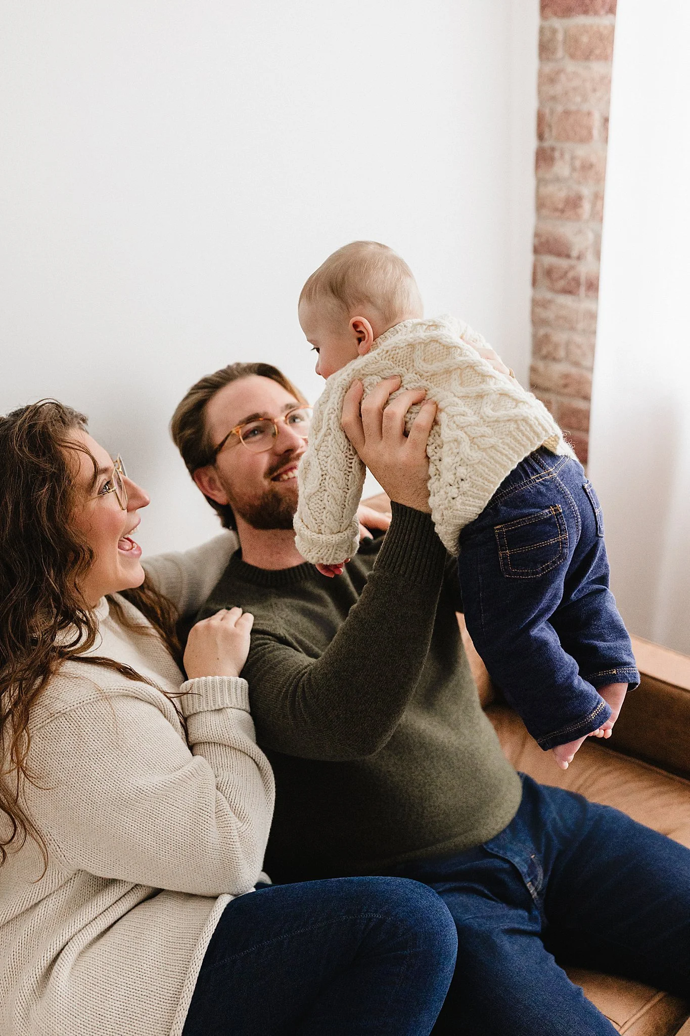 Family of three with a baby, sitting on a couch in a cozy room. The father lifts the baby, who is wearing a cream sweater and jeans, while the mother watches happily during a studio lifestyle family photo session with Ally and B Photography.