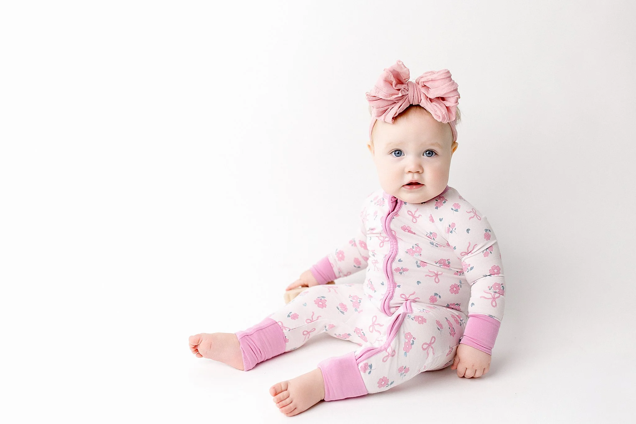 A baby girl with blonde hair and blue eyes sitting on the floor, wearing a pink and white floral onesie with pink cuffs and a pink headband with a bow, against a plain white background for one year old photos near me.