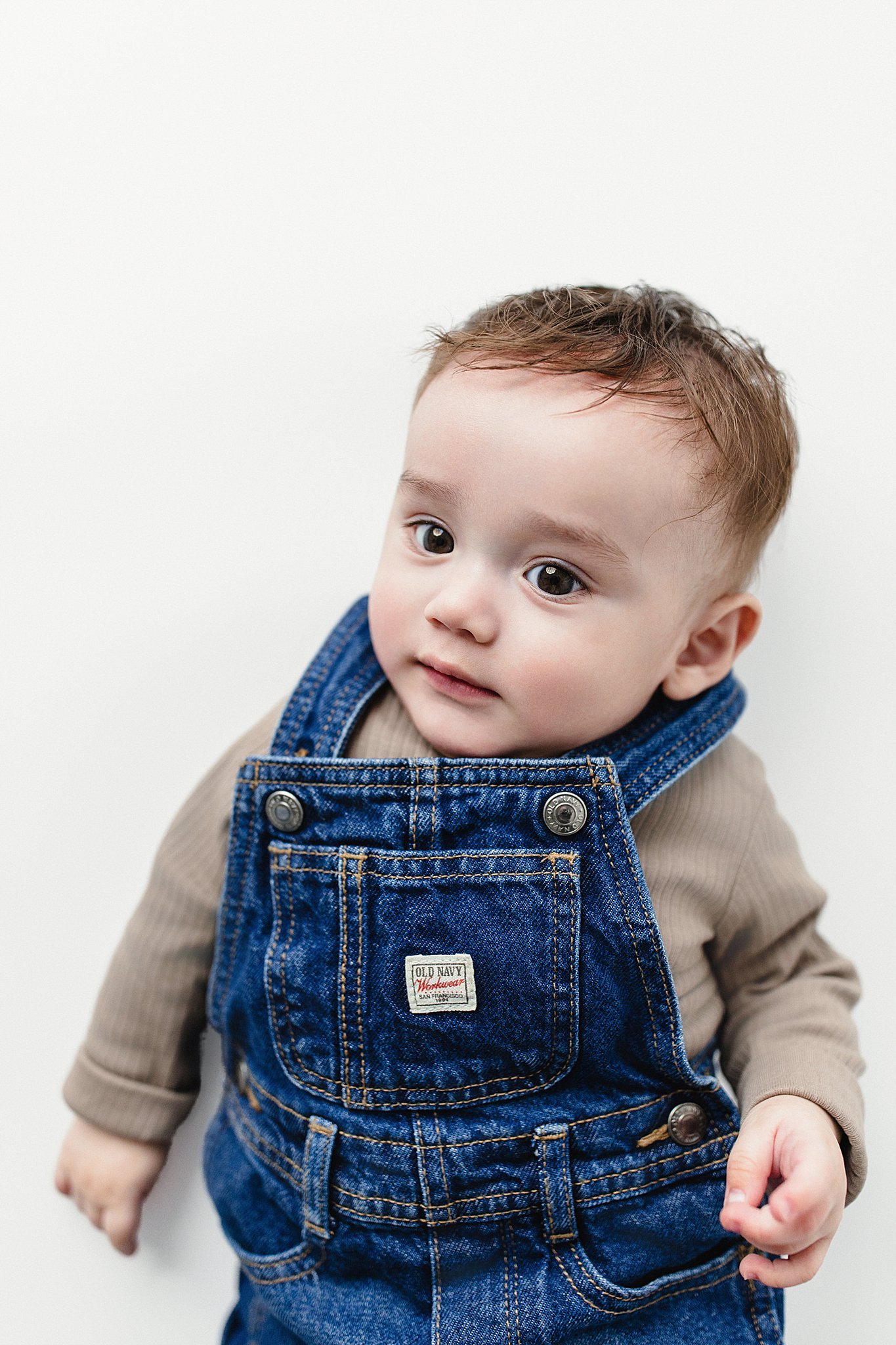 A young child with brown hair and big eyes, wearing a beige shirt and blue denim overalls, looks up at the camera against a plain white background.