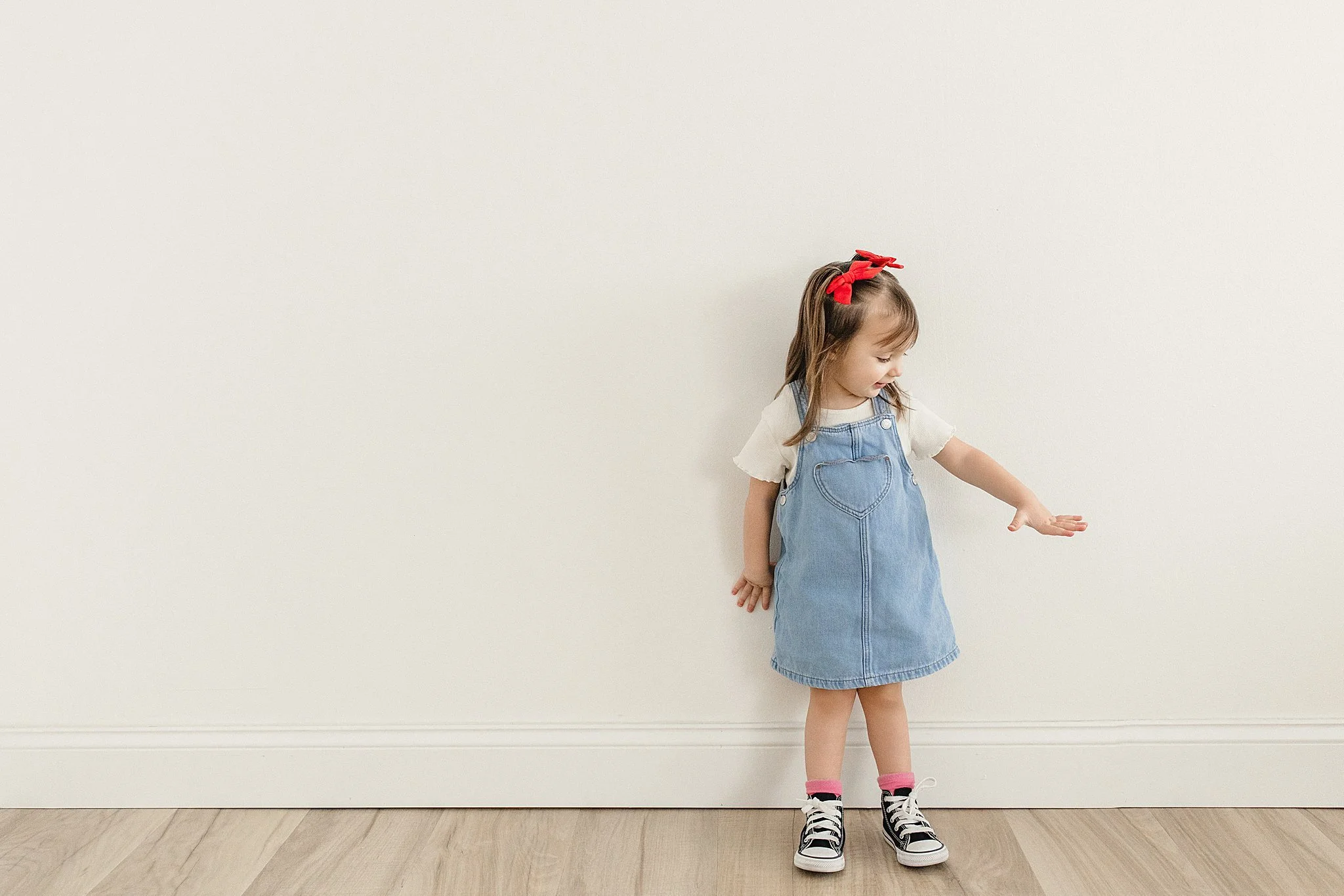 A young girl wearing a denim dress, white t-shirt, pink socks, black sneakers, and a red headband standing against a plain off-white wall, looking and pointing at something during a kids photoshoot in Naperville.