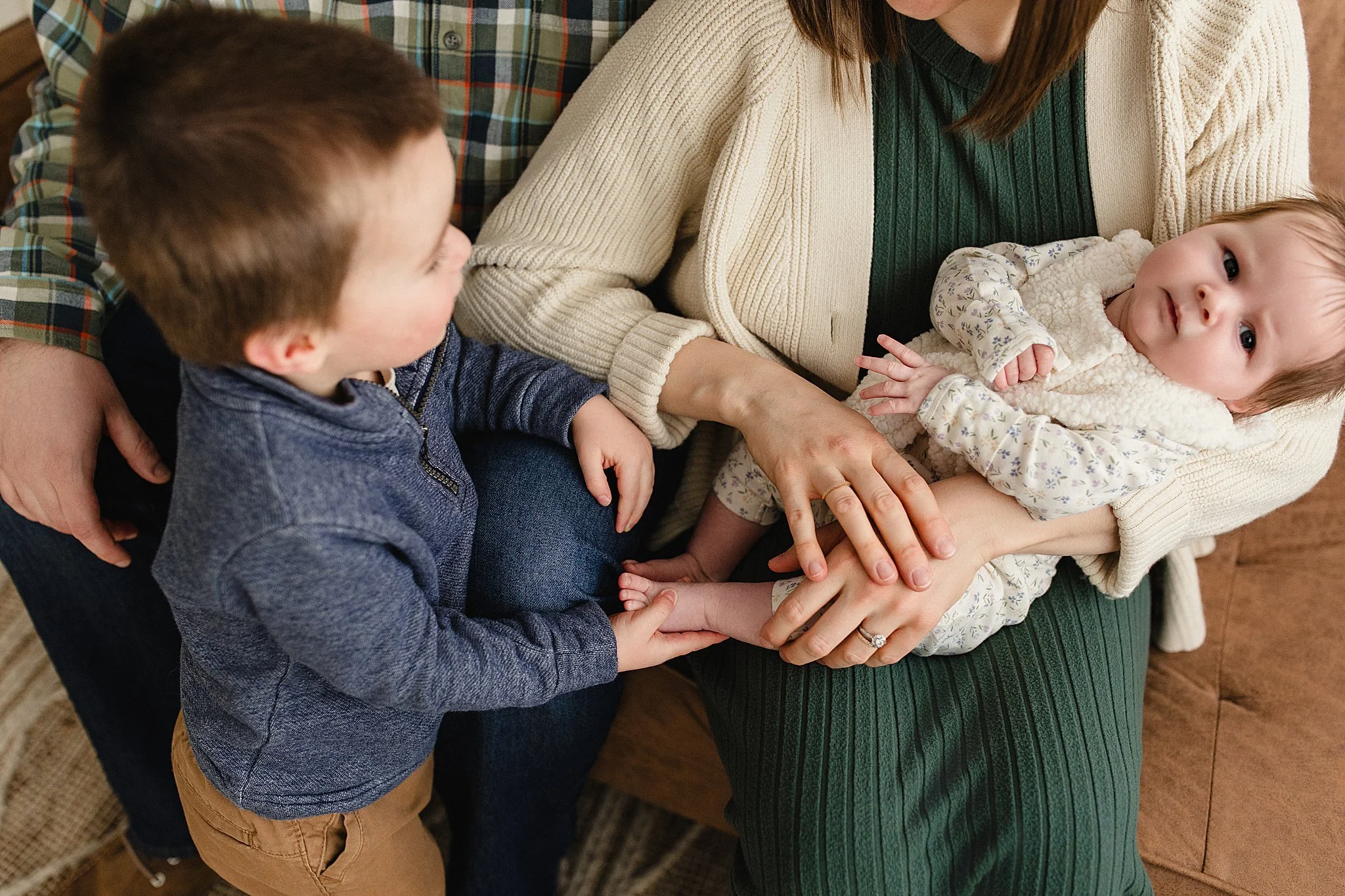 A family scene with a woman holding an infant, a young boy touching the baby's foot, and a man sitting next to them. All are sitting on a couch with the best family photographer Naperville, IL.
