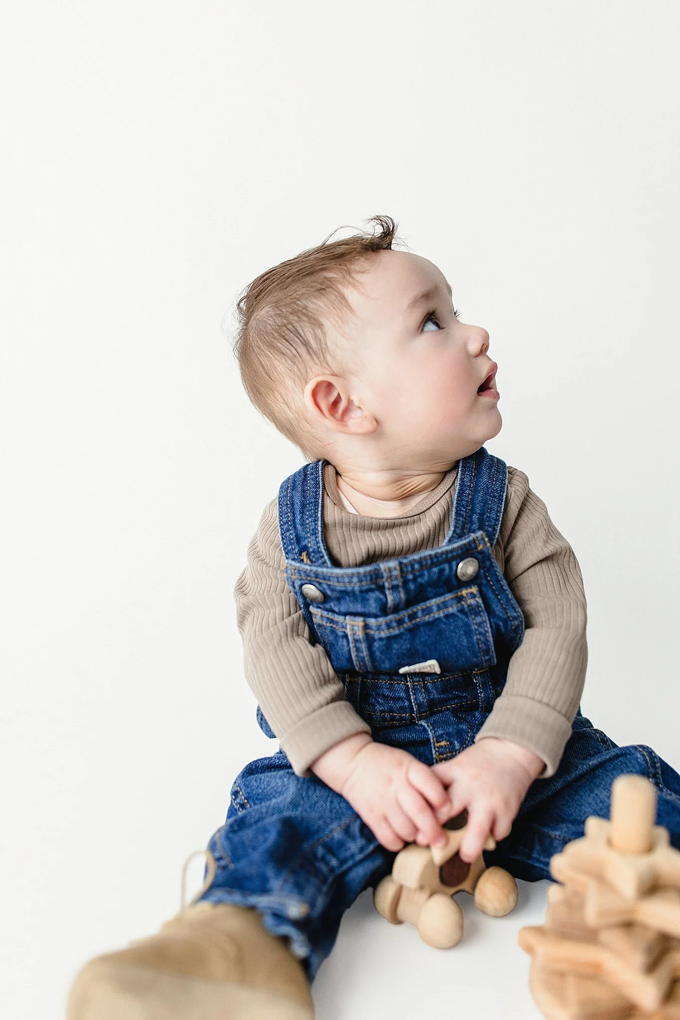 Young boy with reddish hair, wearing tan long sleeves and blue denim overalls, sitting on the floor and looking up, holding wooden building blocks.