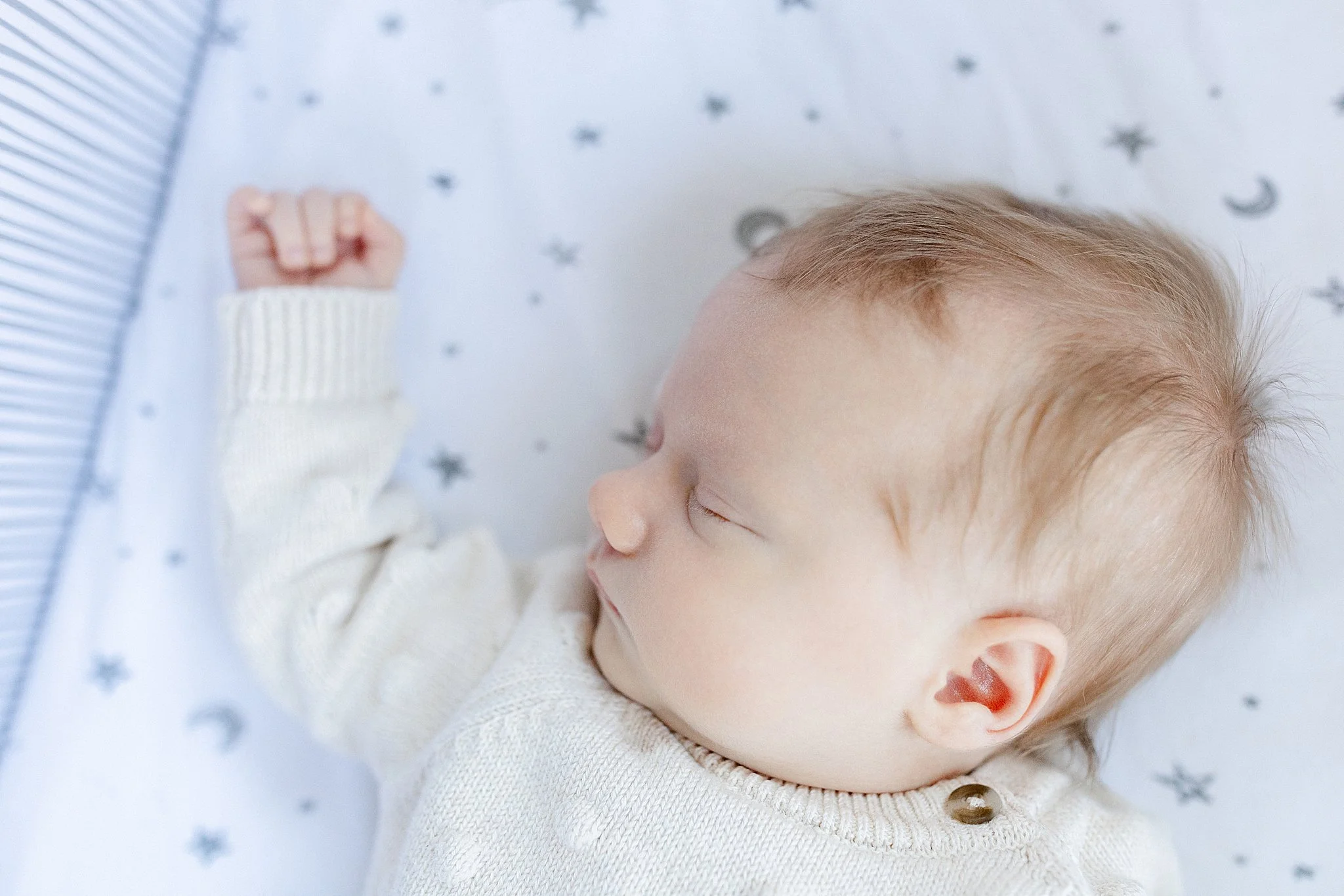 A sleeping baby with blond hair, wearing a white sweater, lying on a star and moon patterned blanket with a lifestyle photographer Plainfield.