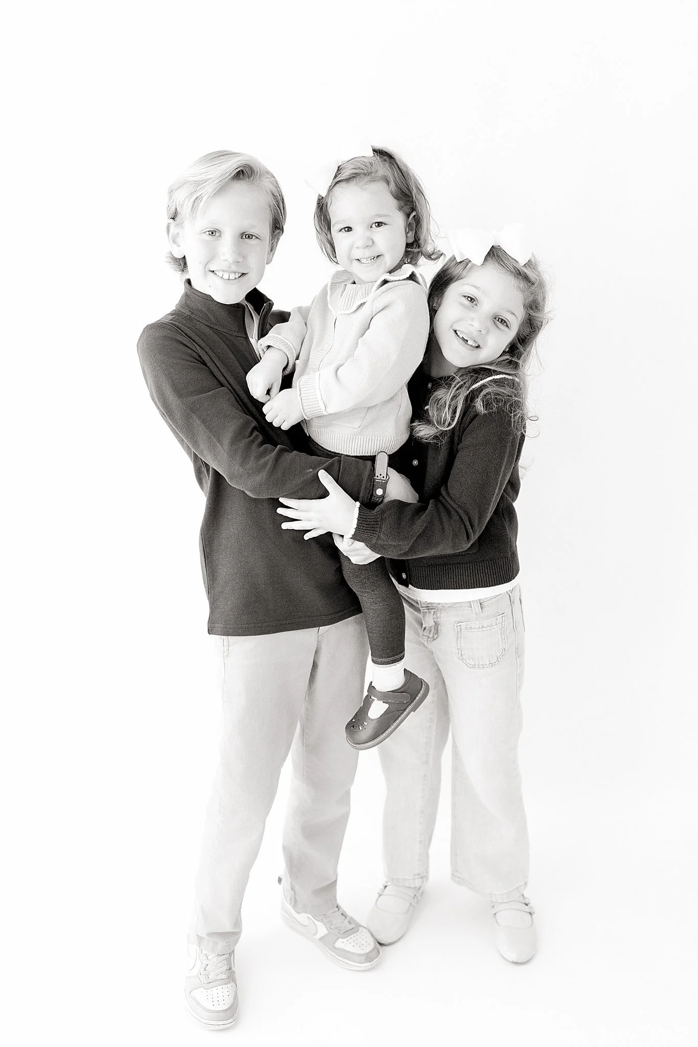A family of three, two children and one adult, posing together and smiling. The adult is holding a young child, and the older child is standing beside them. The image is in black and white during a kids photoshoot Naperville.